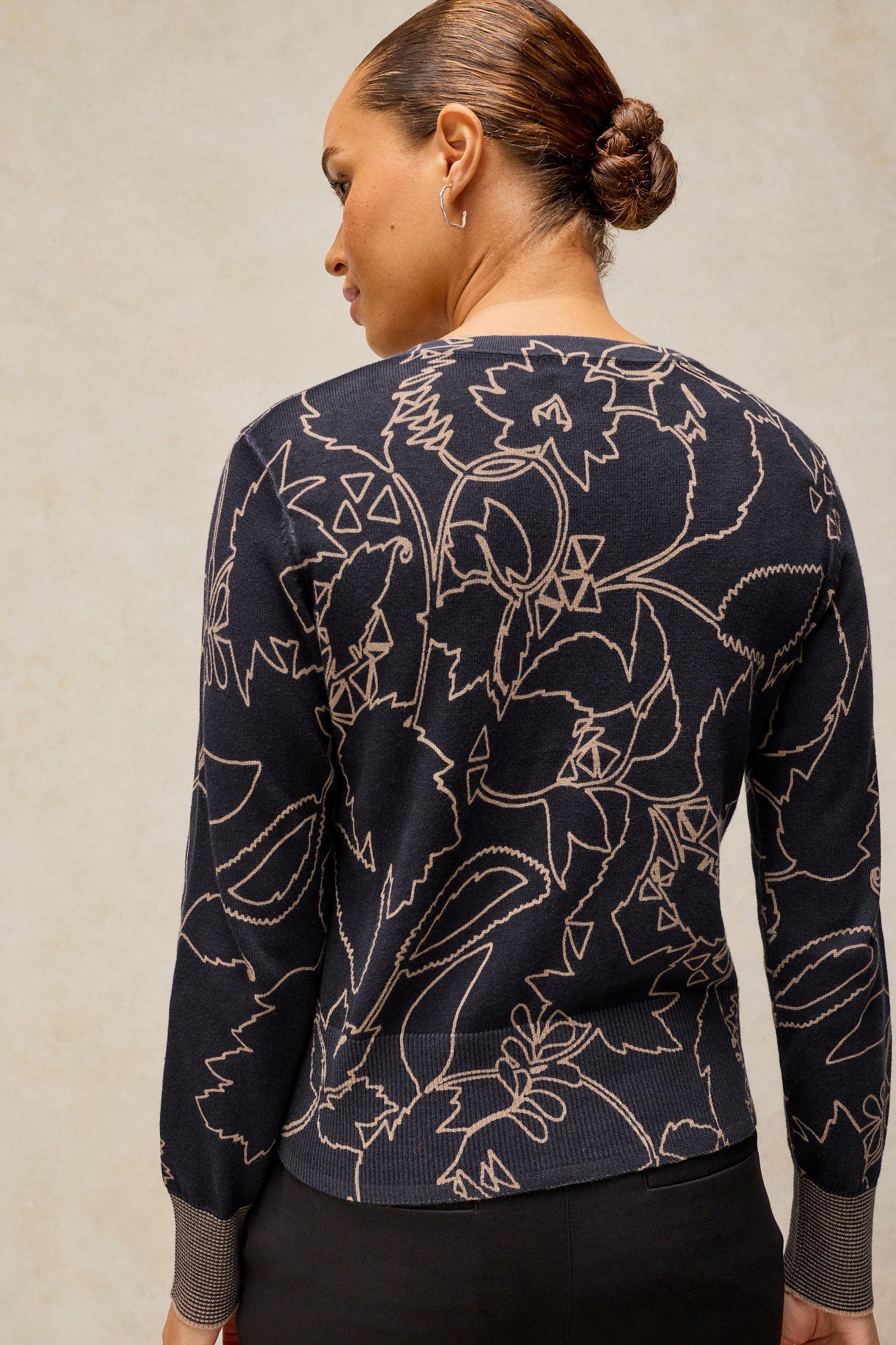 A woman shown from the back wearing a navy blue long-sleeve knit top with a beige floral line pattern and ribbed cuffs, against a neutral textured beige background.