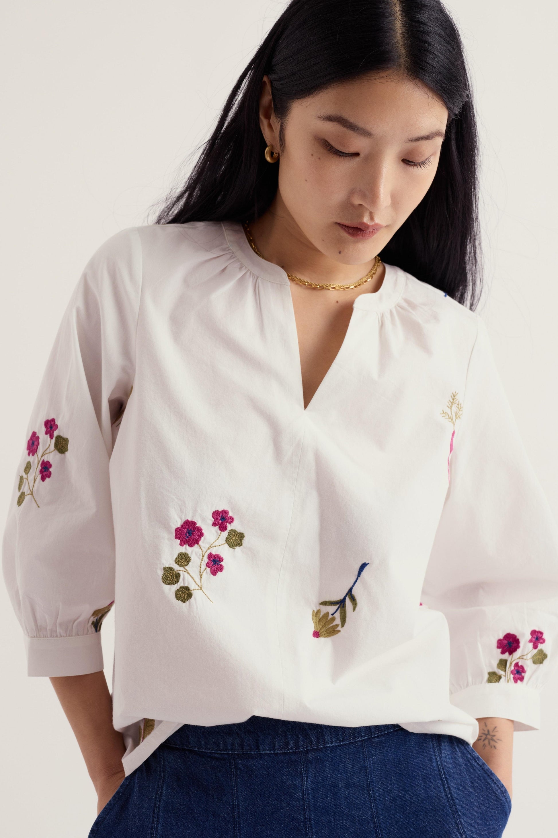 A woman wearing a white blouse with pink and green floral embroidery on the front and sleeves, paired with dark blue jeans, photographed against a light neutral background.