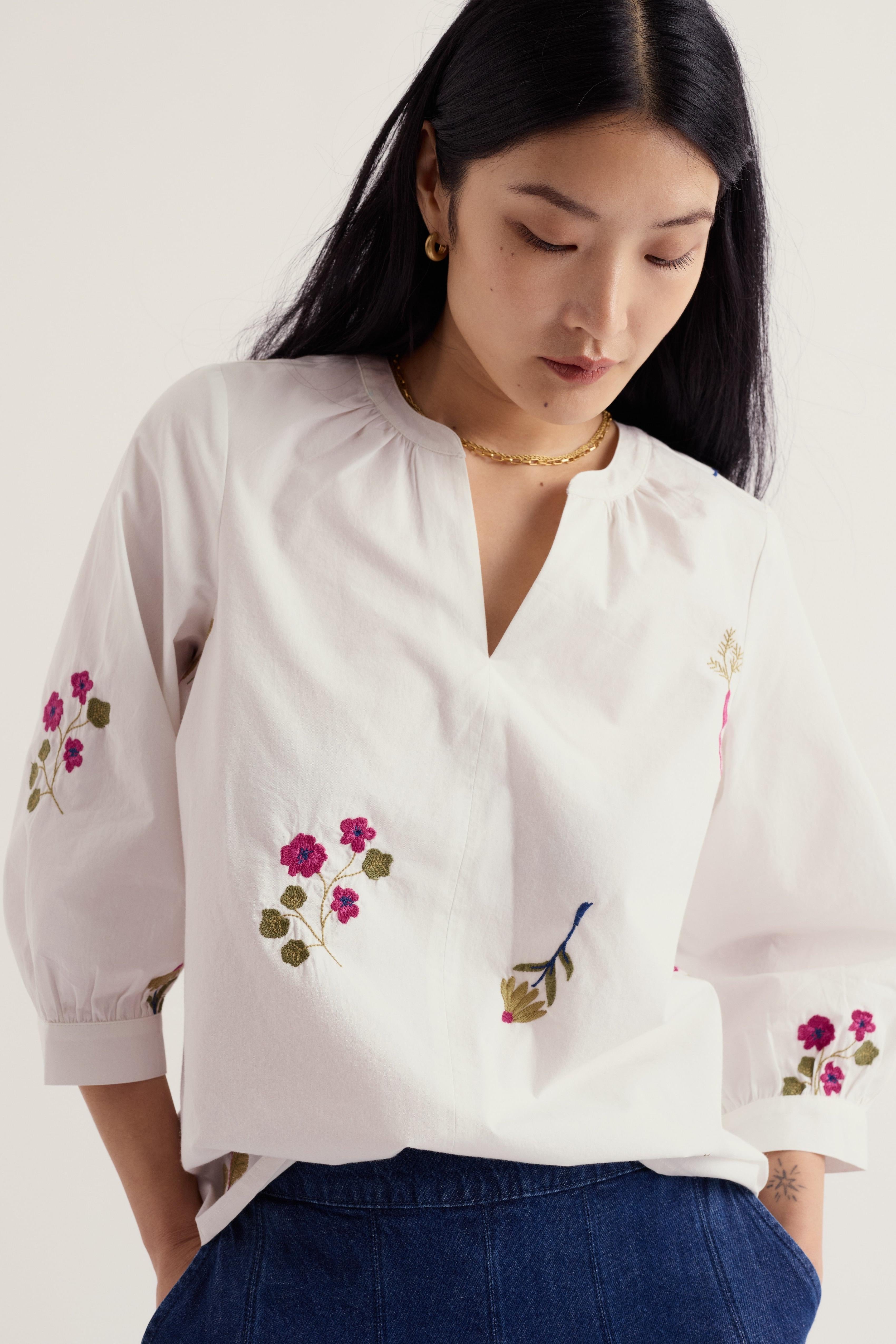 A woman wearing a white blouse with pink and green floral embroidery on the front and sleeves, paired with dark blue jeans, photographed against a light neutral background.