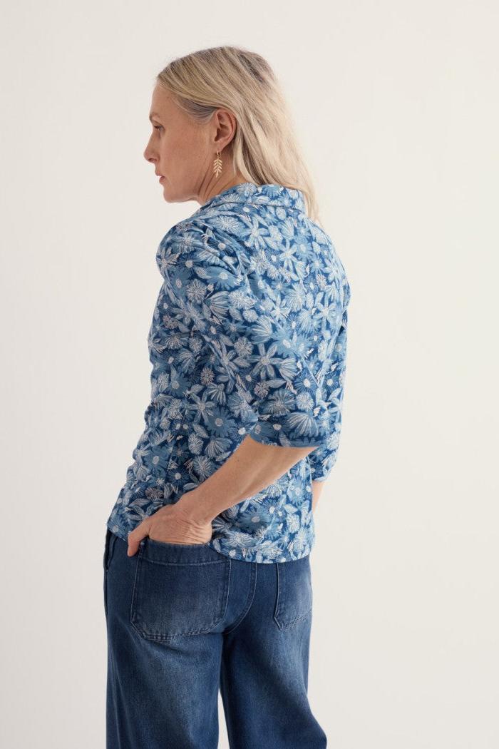 A woman shown from the back wearing a blue floral patterned button-up shirt and dark blue jeans, standing against a plain off-white studio background.