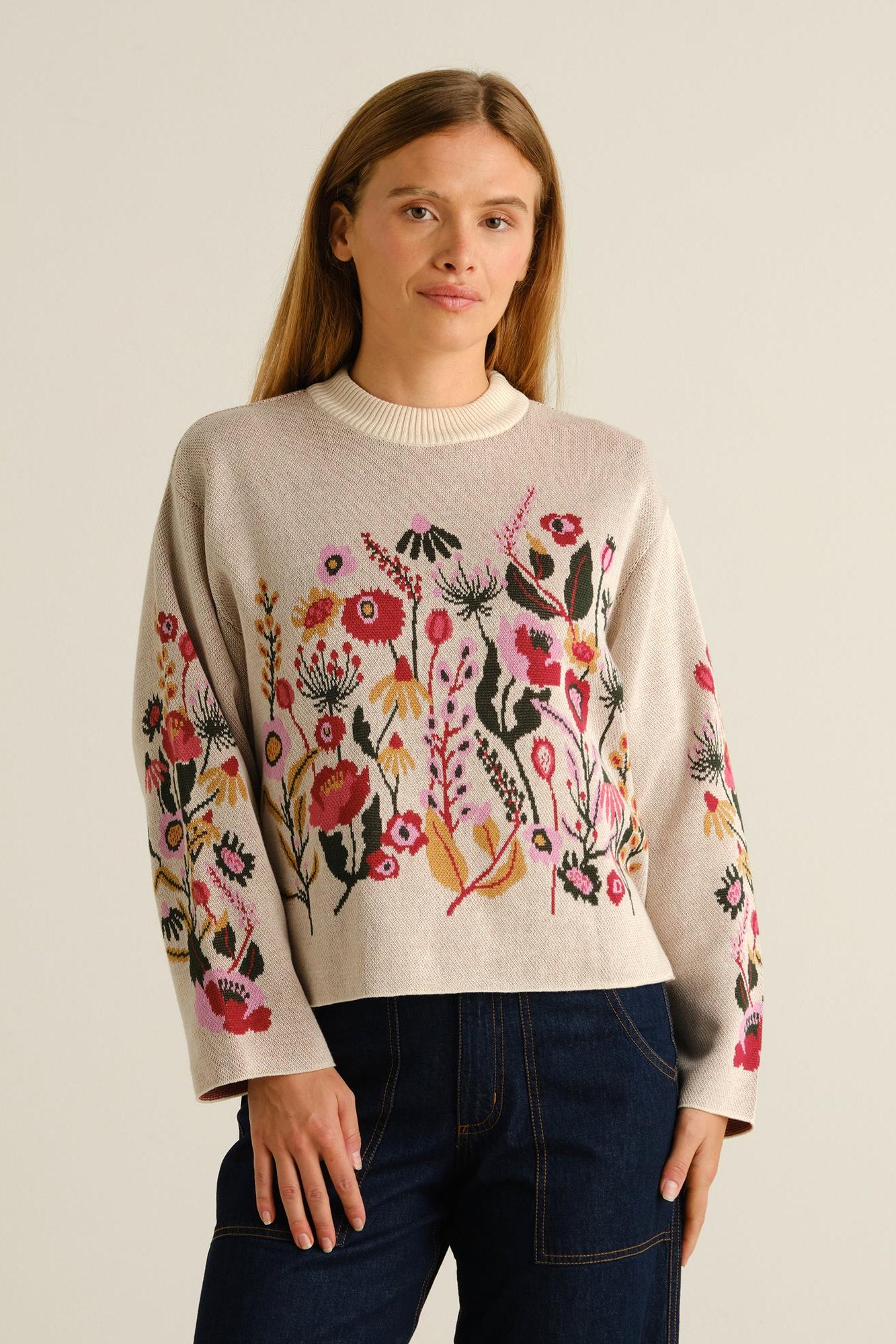 A woman modeling a cream-colored crewneck sweater with vibrant, multicolored floral embroidery across the front and sleeves, paired with dark blue jeans against a plain light beige background.