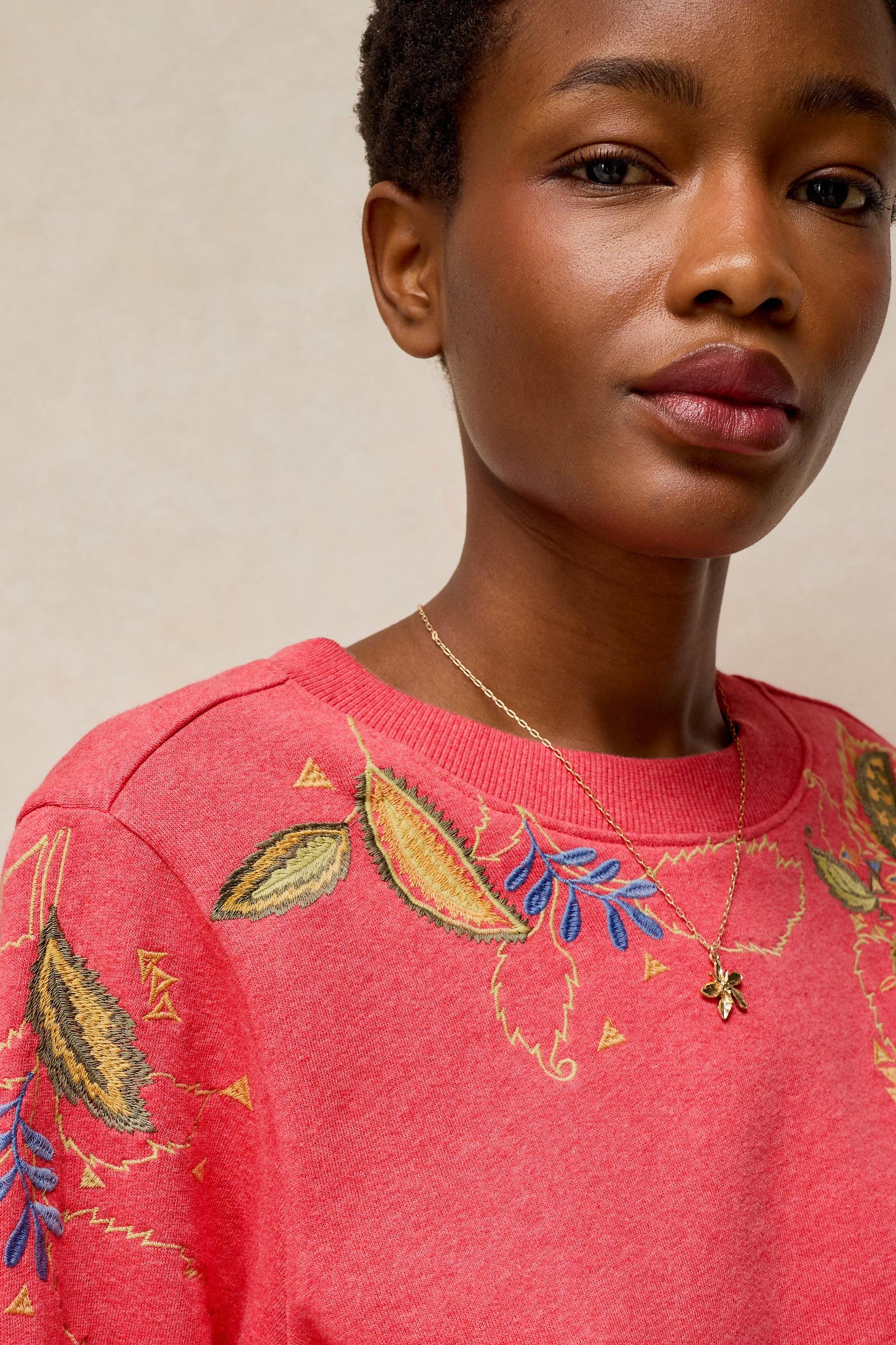 A close-up of a model wearing a coral pink crewneck sweatshirt with embroidered botanical leaf patterns in gold, blue, and green.
