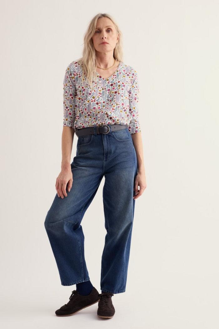 A woman modeling a floral print blouse with elbow-length sleeves and dark blue wide-leg jeans, paired with a black belt and dark brown shoes, against a plain light background.