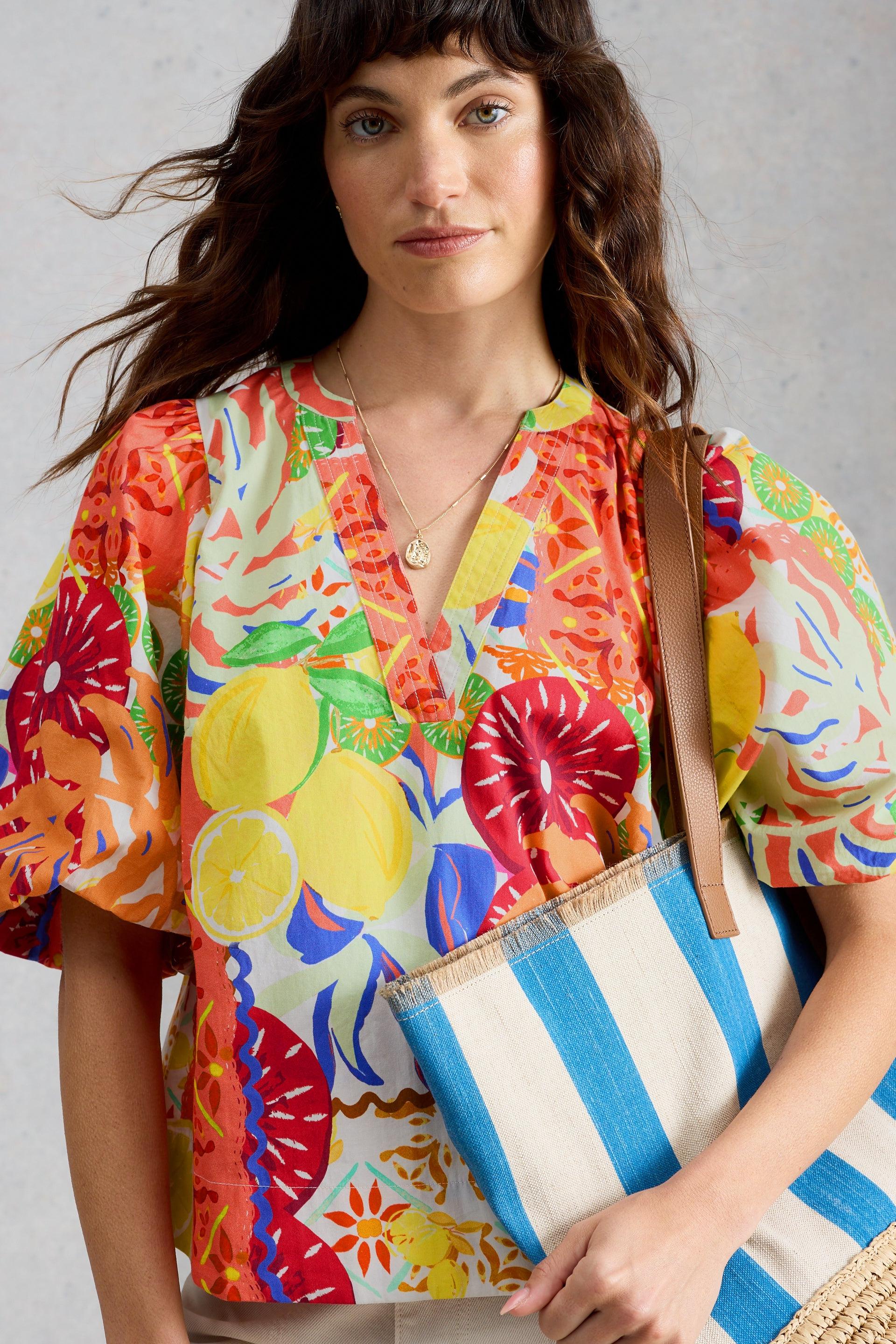 Woman wearing a colourful fruit-print organic cotton top by White Stuff while carrying a striped straw bag.