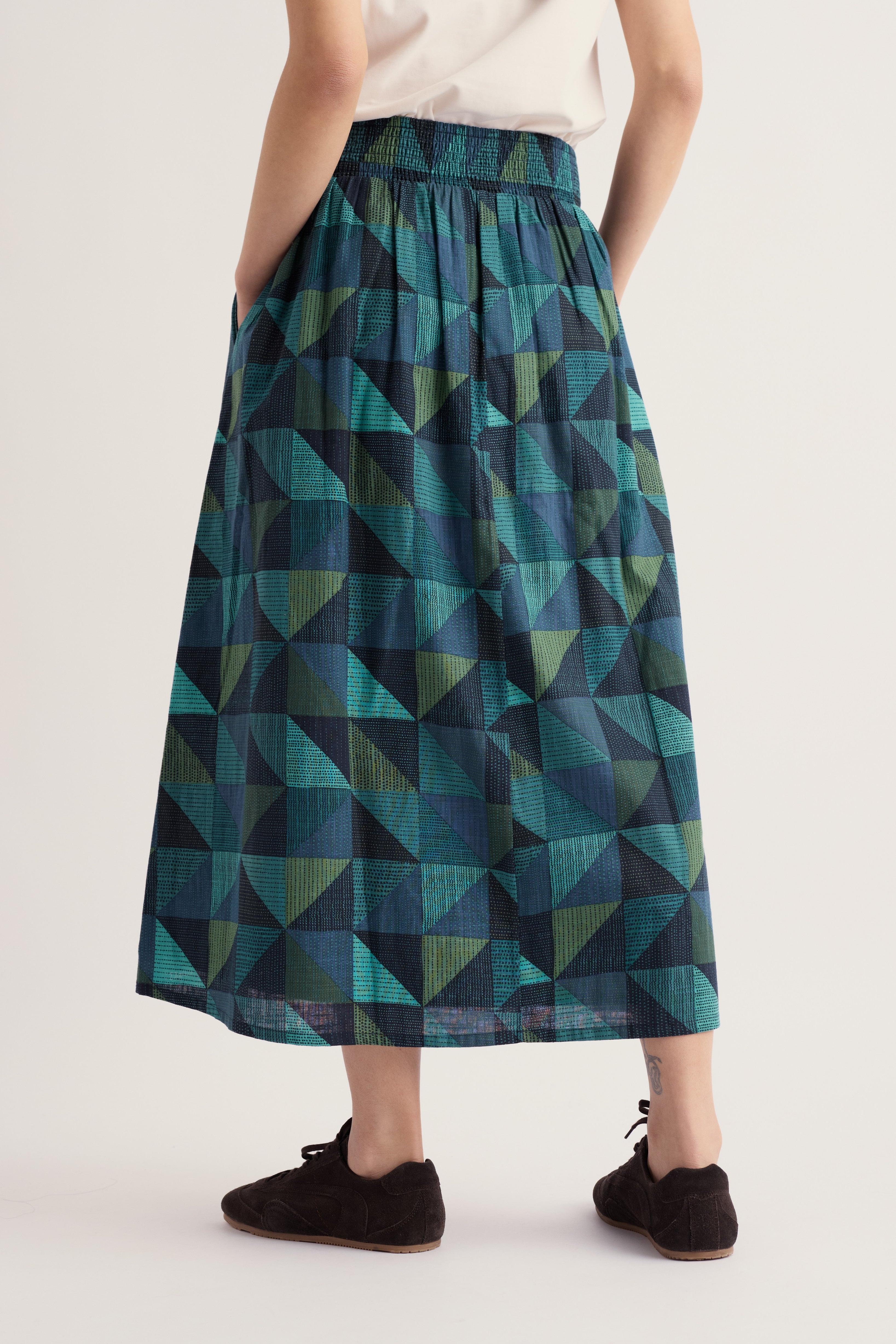 A person wearing a high-waisted midi skirt with a geometric triangle pattern in teal, green, and dark blue, paired with a white top and black slip-on shoes, shown from the waist down against a light gray studio background.