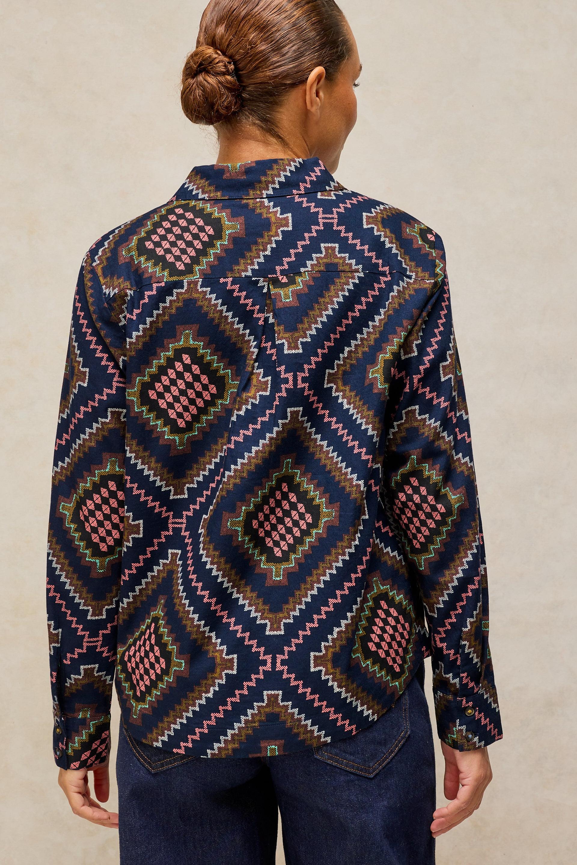 A woman shown from the back wearing a long-sleeve button-up shirt with a complex geometric diamond pattern in navy blue, pink, olive green, and brown, paired with dark blue jeans against a neutral beige background.