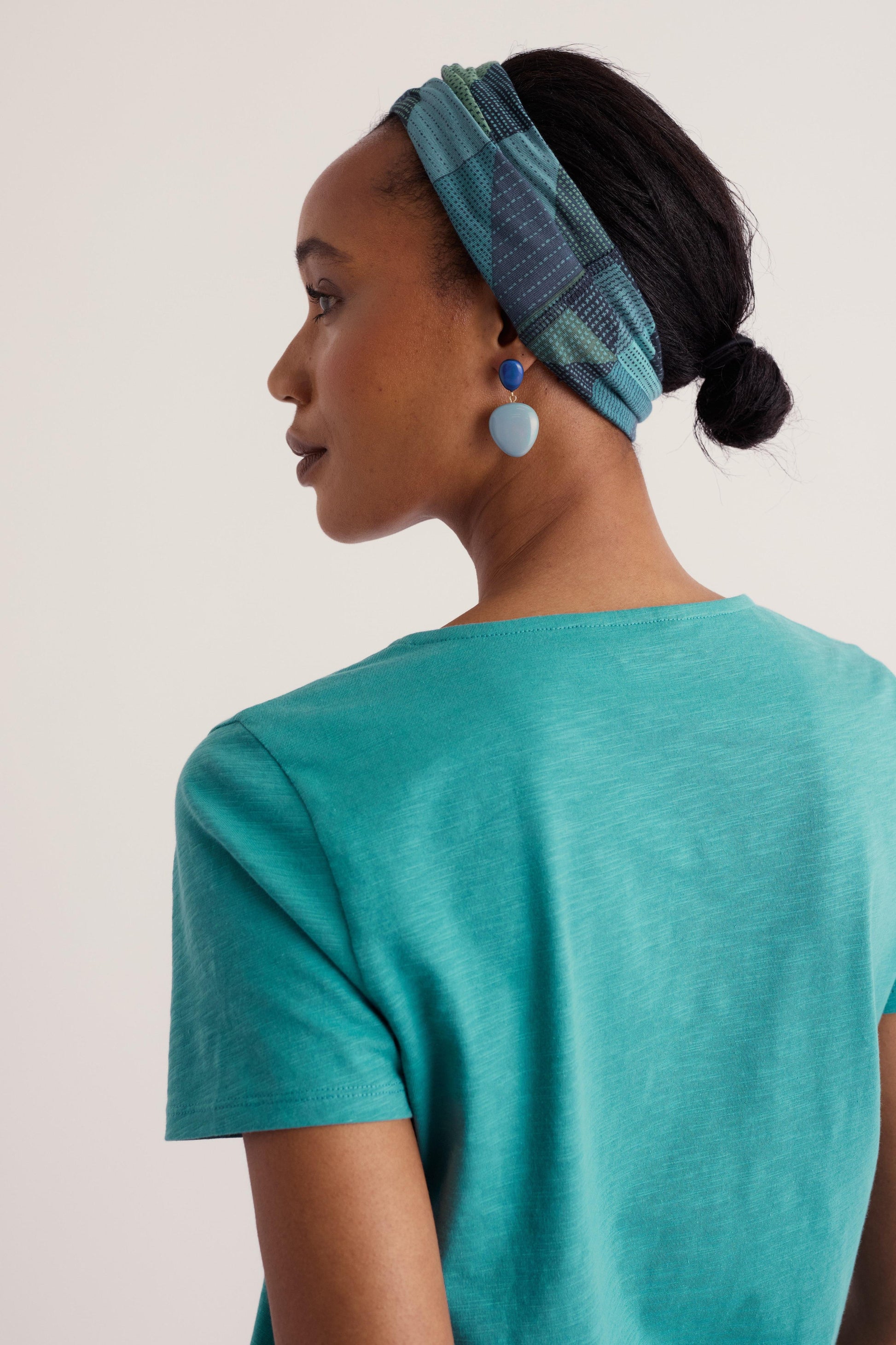 A woman shown from the back wearing a teal short-sleeve top and a blue plaid headband, against a plain light beige studio background.