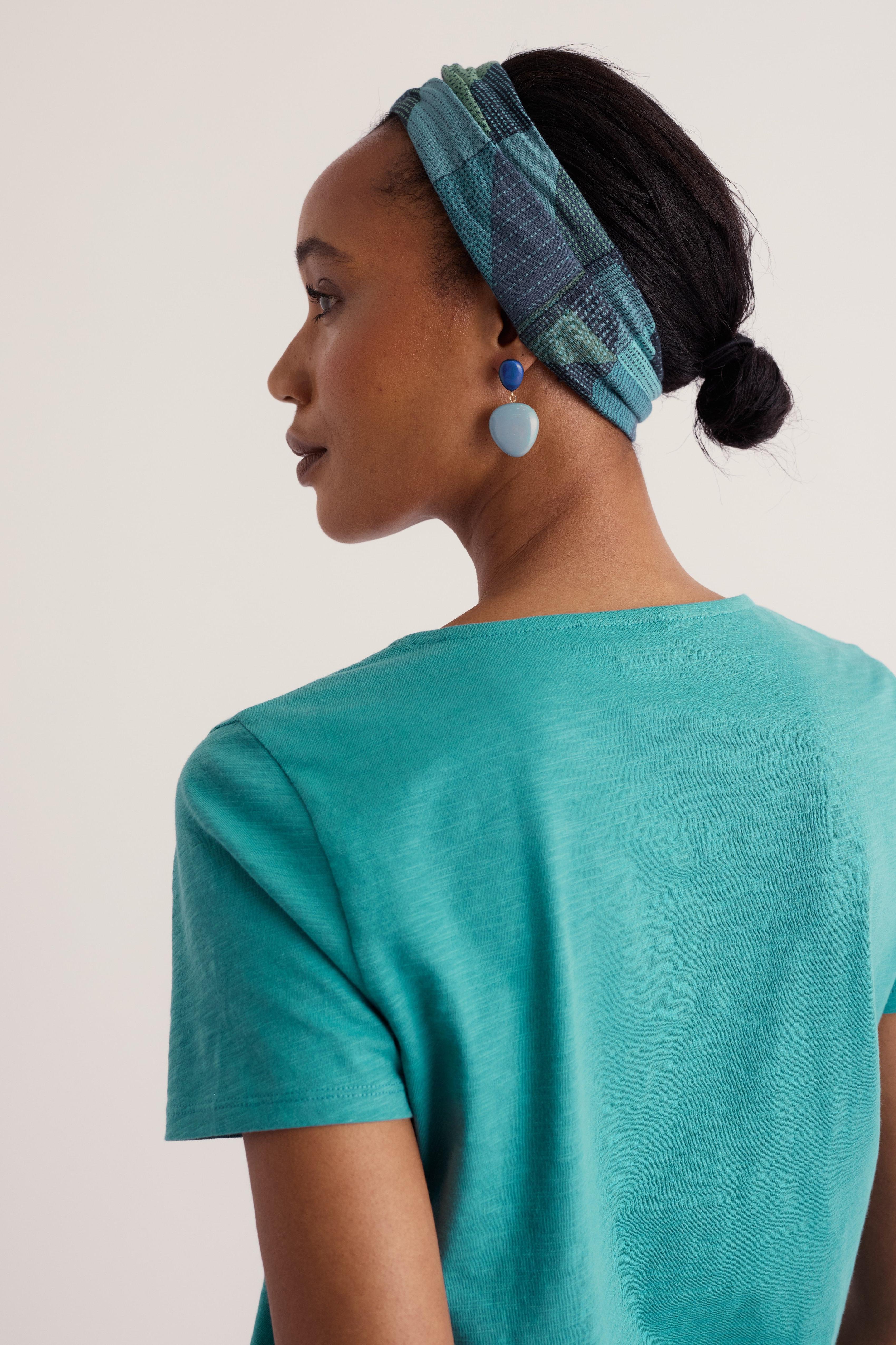 A woman shown from the back wearing a teal short-sleeve top and a blue plaid headband, against a plain light beige studio background.
