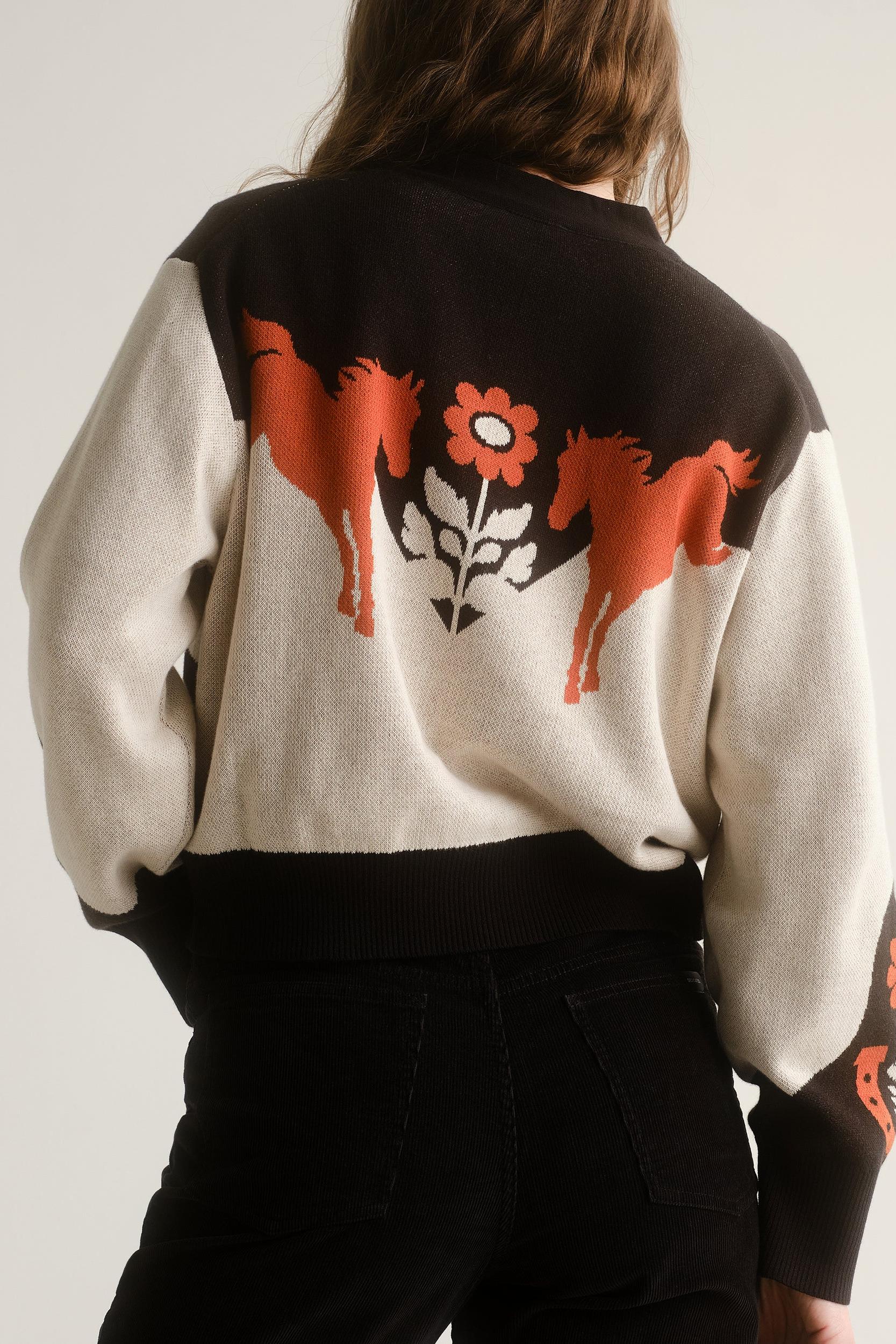 A person shown from the back wearing a knit sweater with a cream body, black shoulders and hem, and orange horse and flower graphics across the upper back.