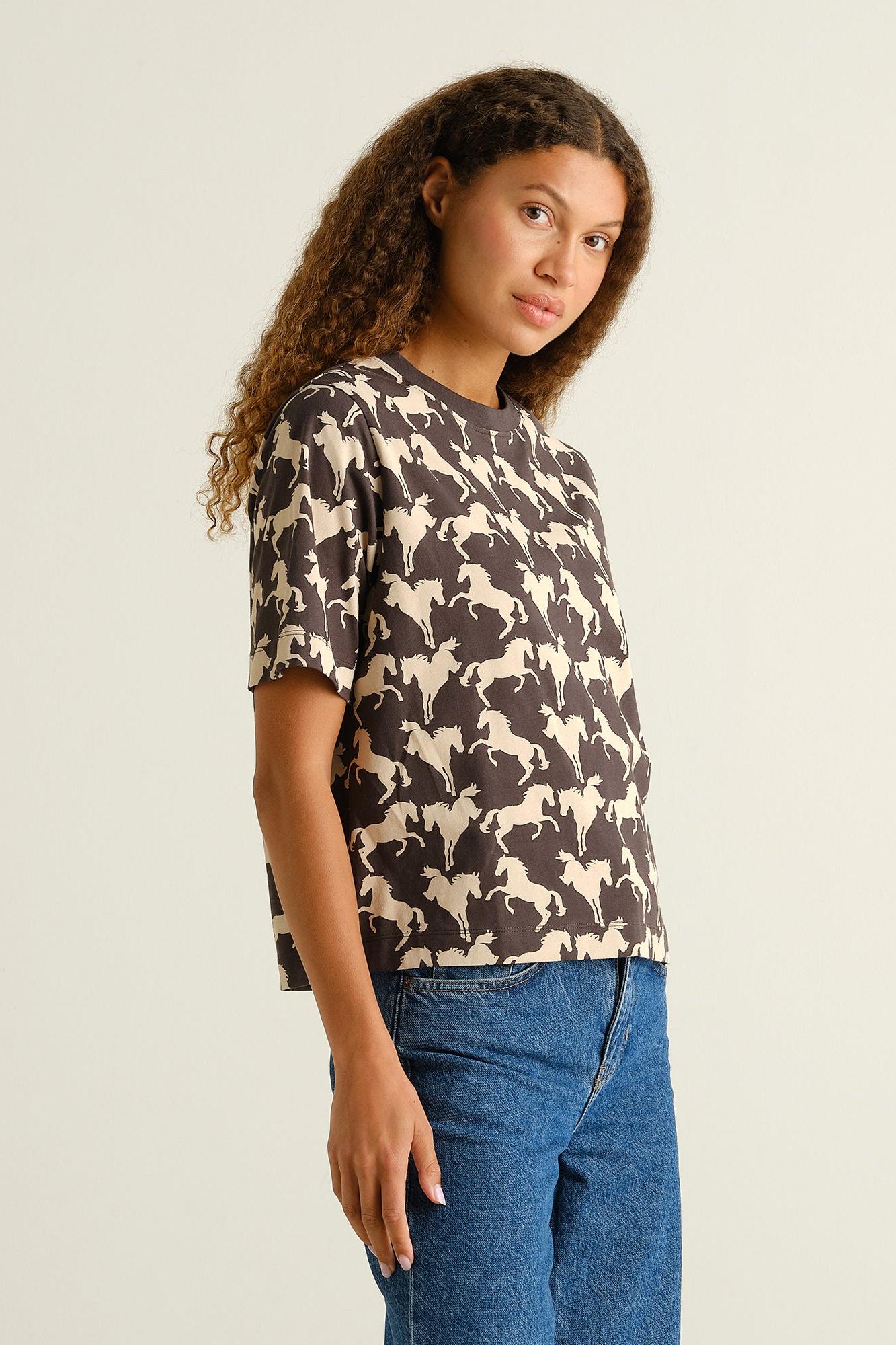 A woman modeling a short-sleeve crewneck t-shirt with an all-over horse pattern in dark brown and cream, paired with blue jeans, against a plain light beige background.