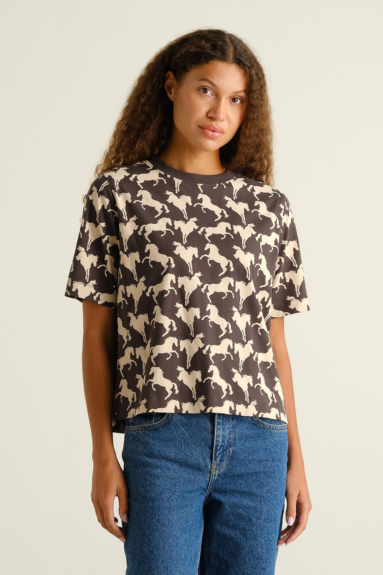 A woman modeling a dark brown short-sleeve t-shirt with an all-over cream horse pattern, paired with blue jeans, against a plain light beige background.