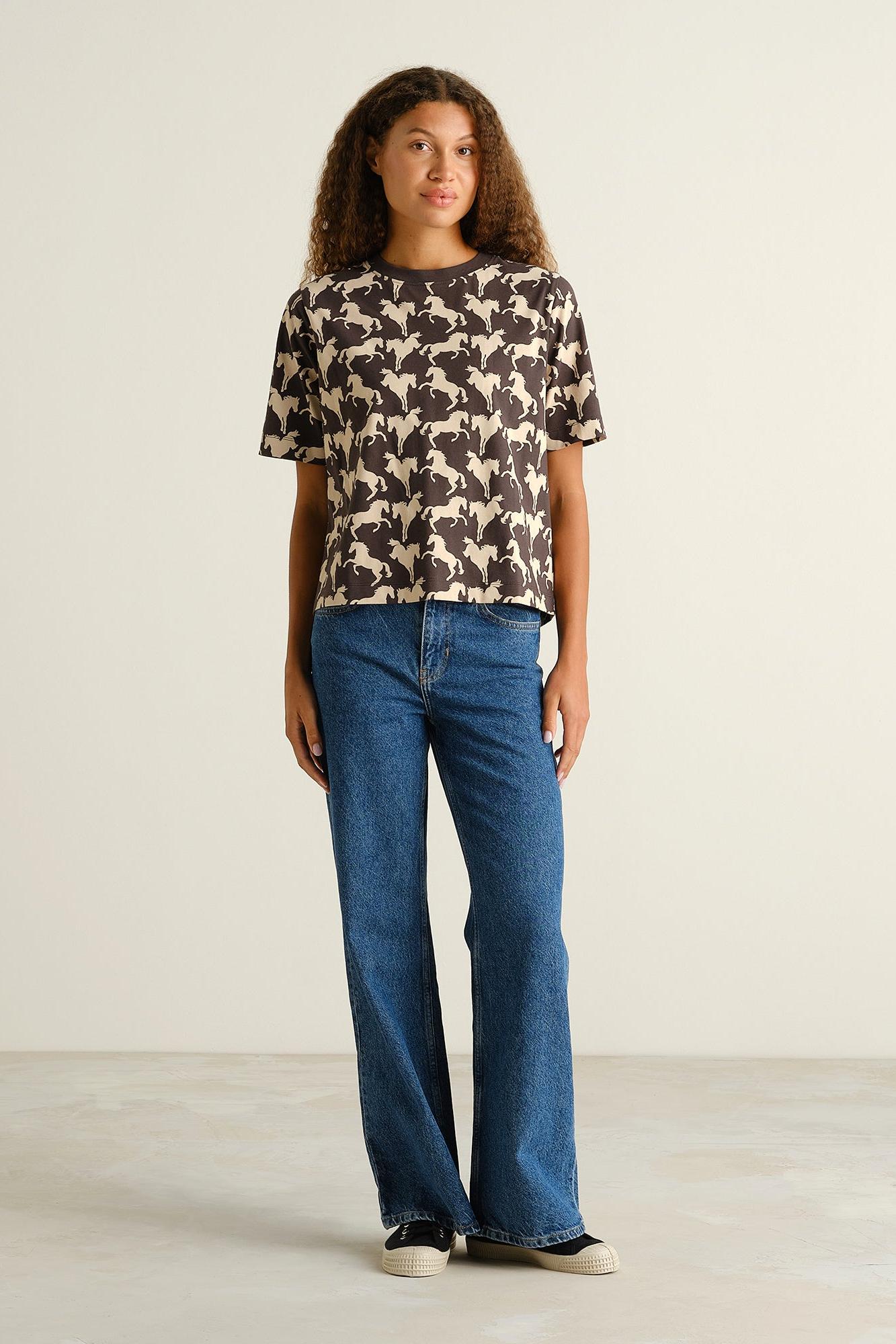 A woman stands in a studio wearing a short-sleeve crewneck top with a beige and black animal print, paired with blue jeans and black sneakers.