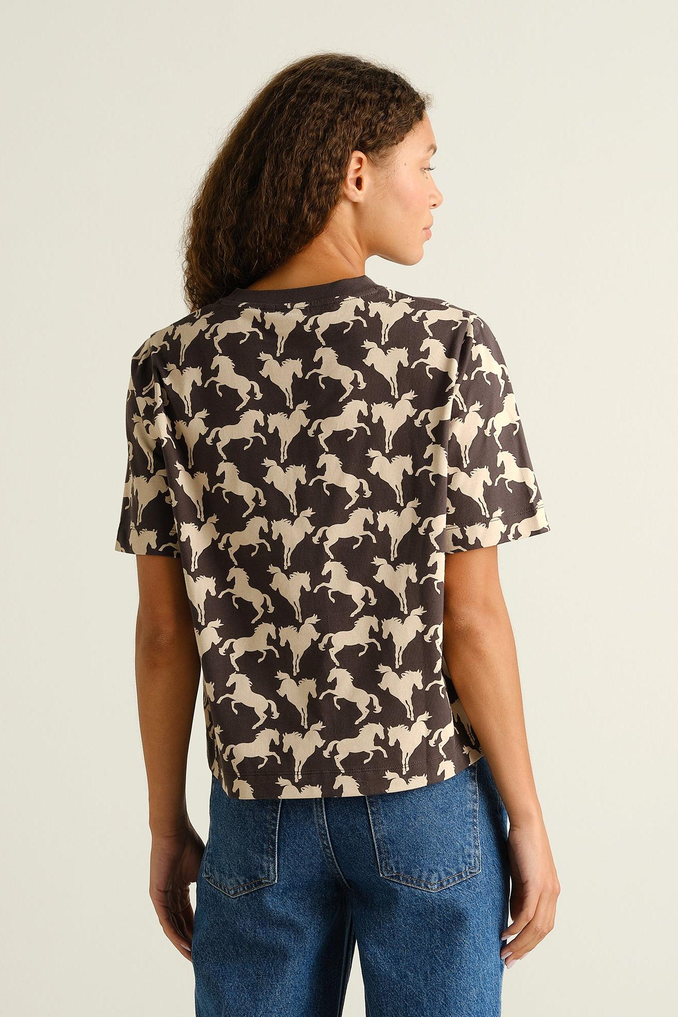 A woman shown from the back wearing a short-sleeve t-shirt with an all-over beige and dark brown horse pattern, paired with blue jeans, against a plain light background.