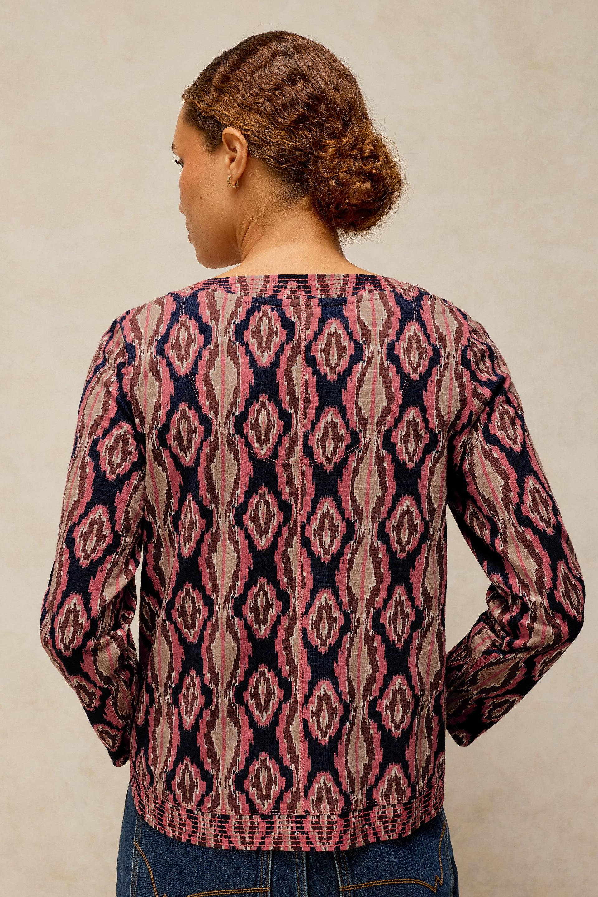 A woman viewed from the back wearing a long-sleeve top with a pink, navy, and beige geometric ikat pattern, paired with blue jeans against a neutral beige background.