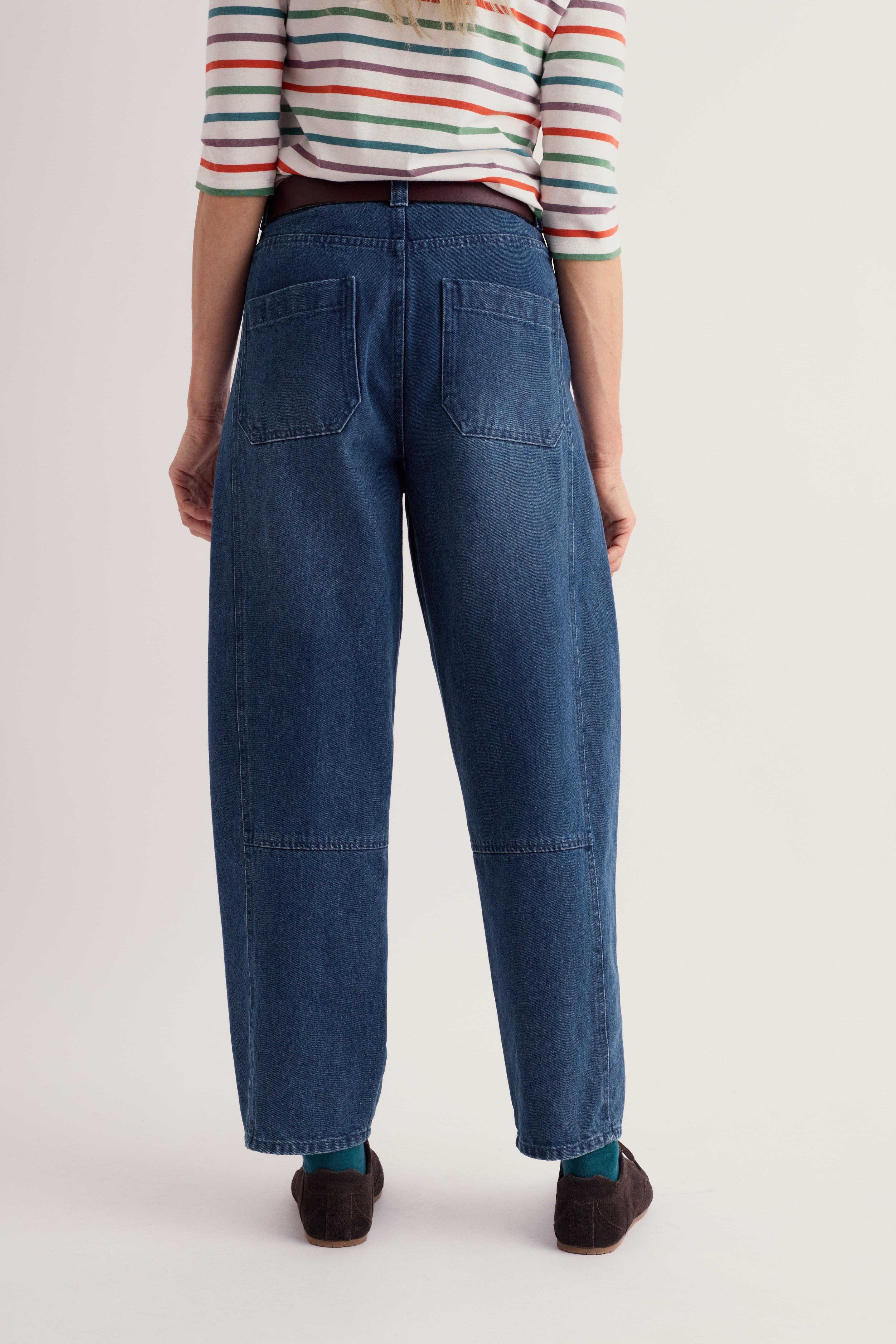 Rear view of a person wearing loose-fit blue denim jeans with patch pockets, a striped long-sleeve top, and dark slip-on shoes against a plain light background.
