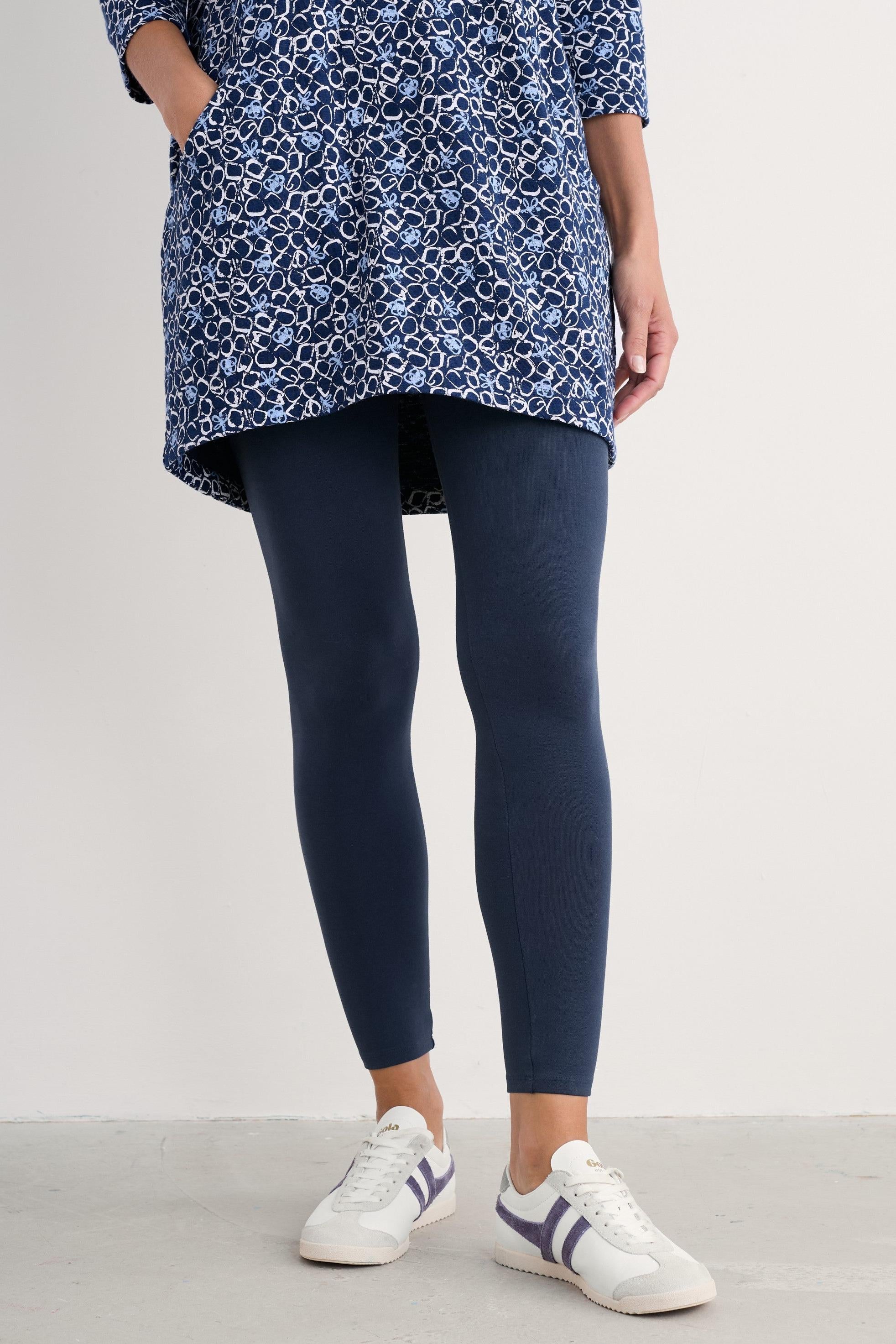 A model from the waist down wearing a blue and white patterned tunic top, navy blue leggings, and white sneakers with blue stripes, standing against a plain light gray wall.