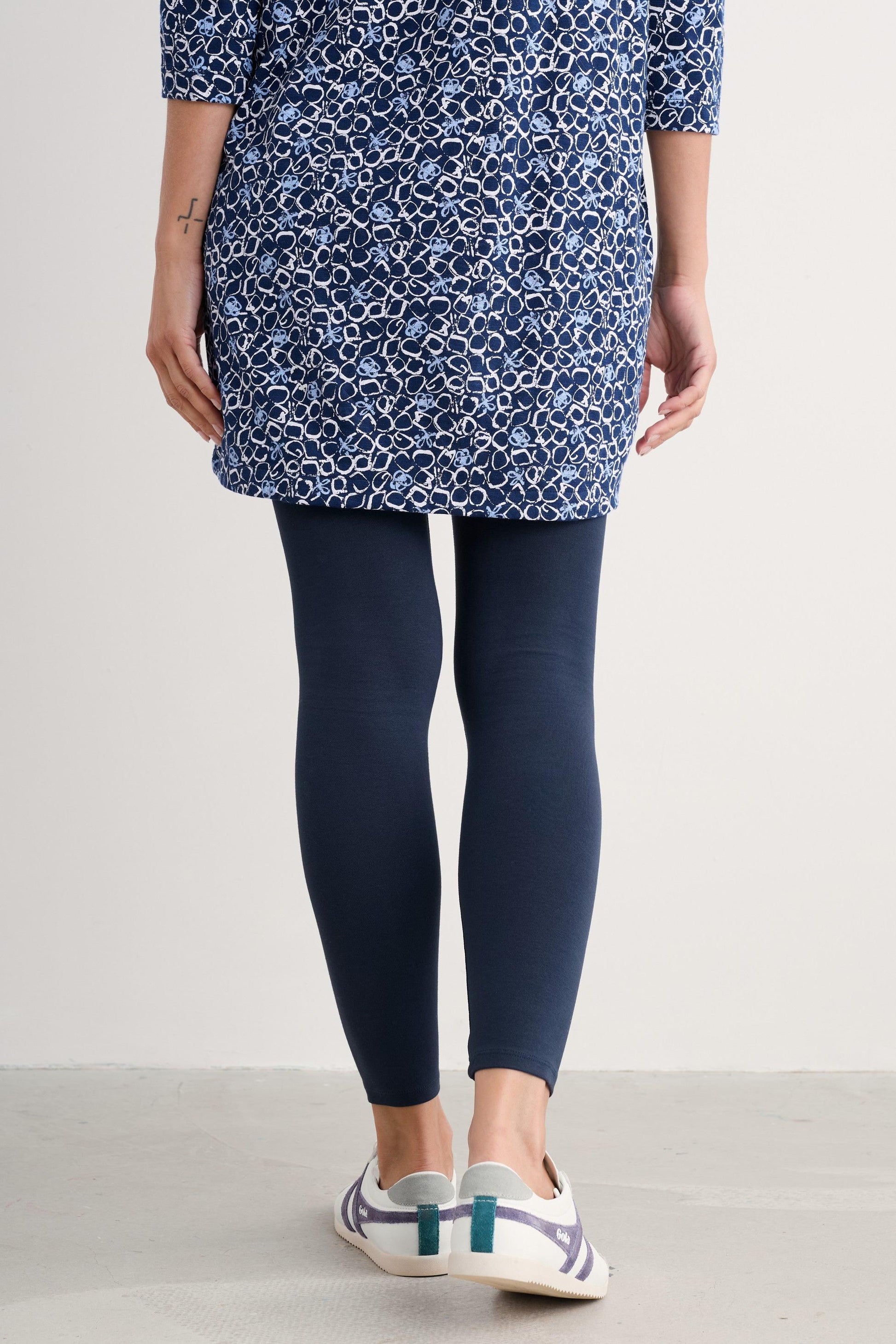 A model is shown from the waist down wearing a blue and white patterned tunic top, dark navy leggings, and white sneakers with teal accents, set against a plain light gray studio background.