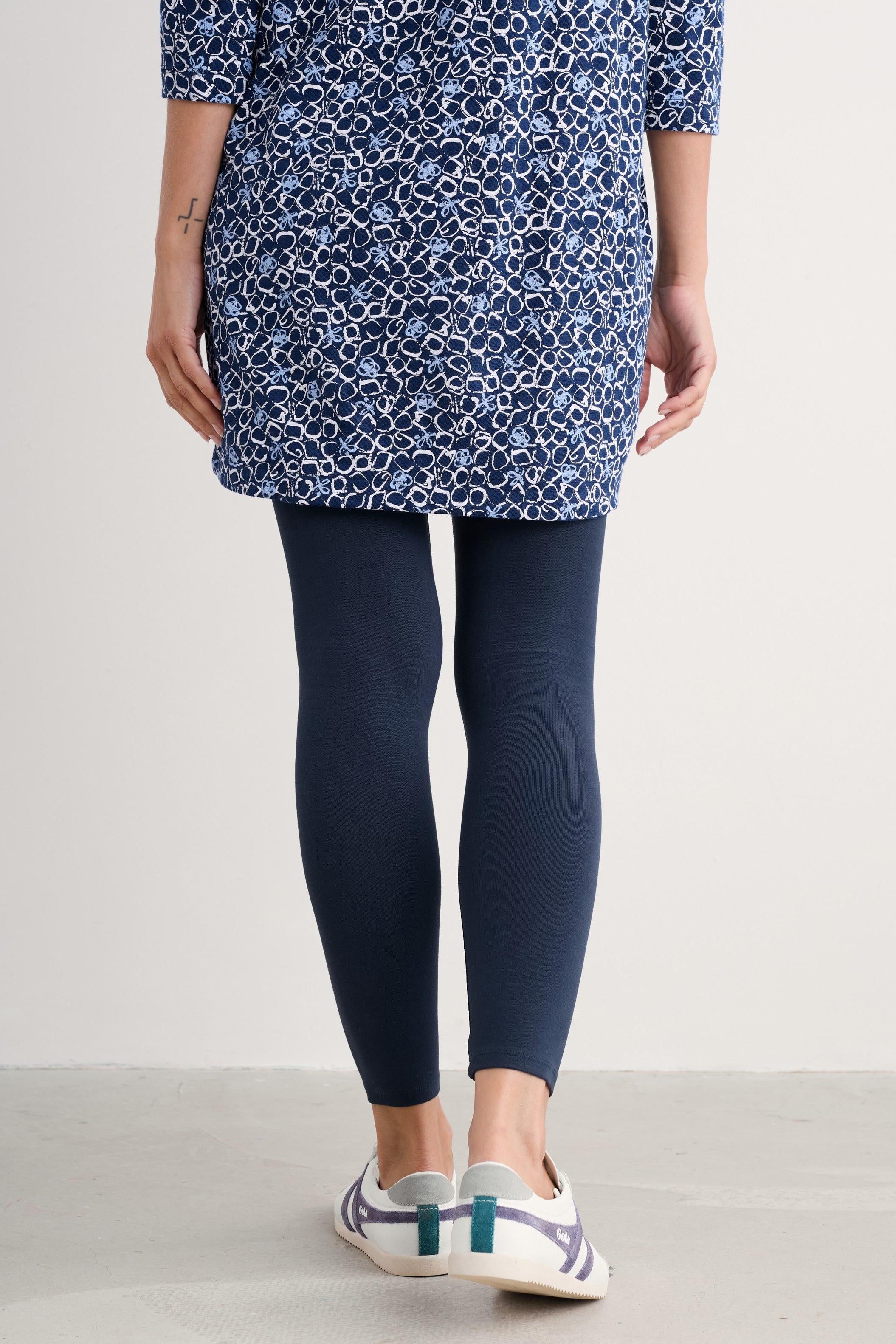 A model is shown from the waist down wearing a blue and white patterned tunic top, dark navy leggings, and white sneakers with teal accents, set against a plain light gray studio background.