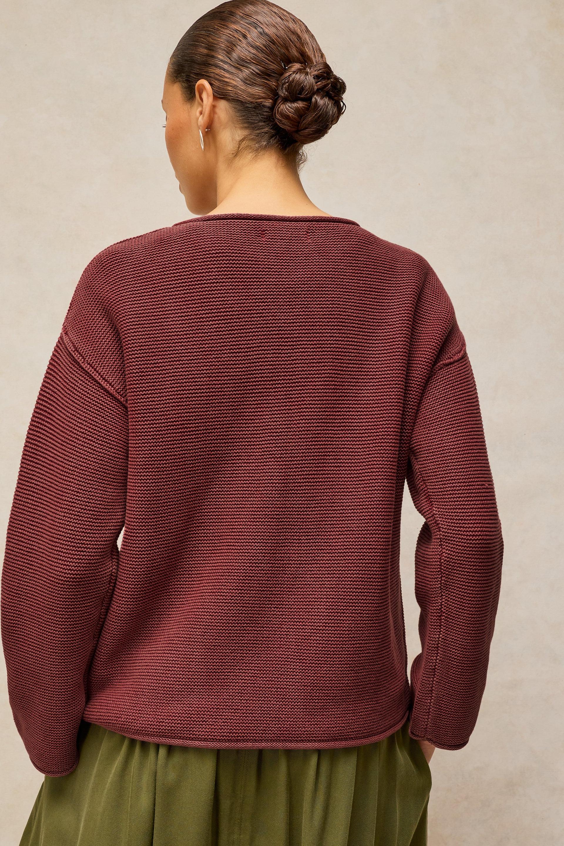 A woman shown from the back wearing a ribbed knit sweater in a deep burgundy color, paired with an olive green skirt, against a textured beige background.