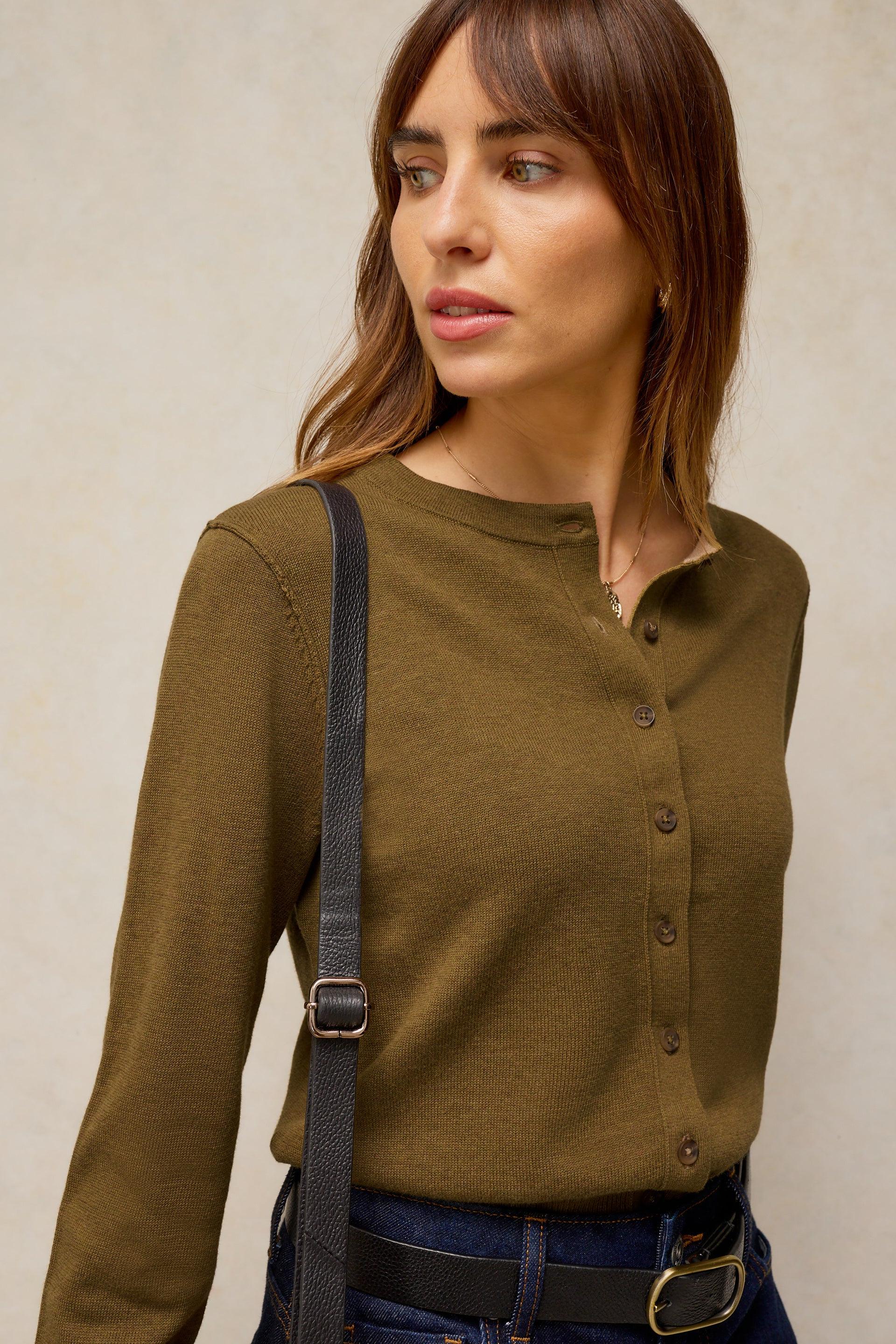 A woman wearing a long-sleeve olive green henley-style top with a buttoned front, paired with dark blue jeans and a black crossbody bag strap, against a neutral textured beige background.