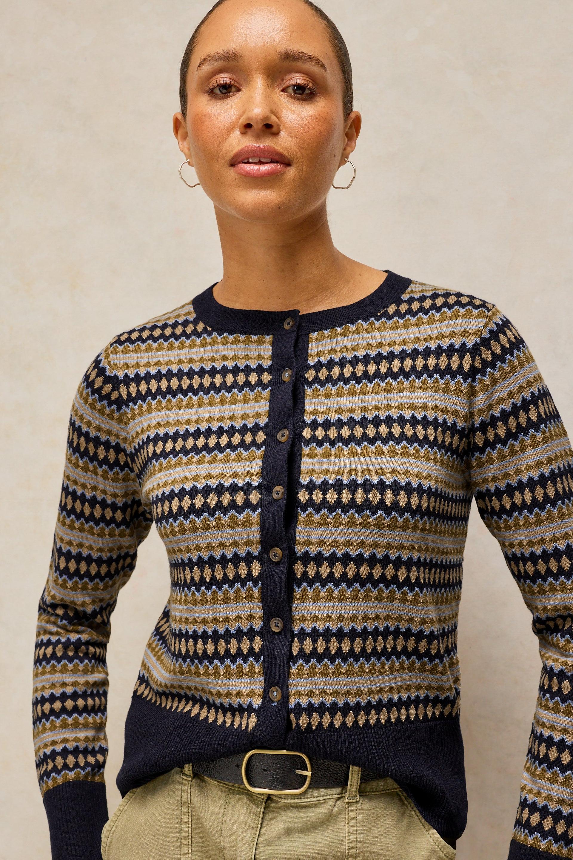 A woman wearing a navy, beige, and gold patterned button-up cardigan sweater, paired with khaki pants and a belt, against a neutral textured background.