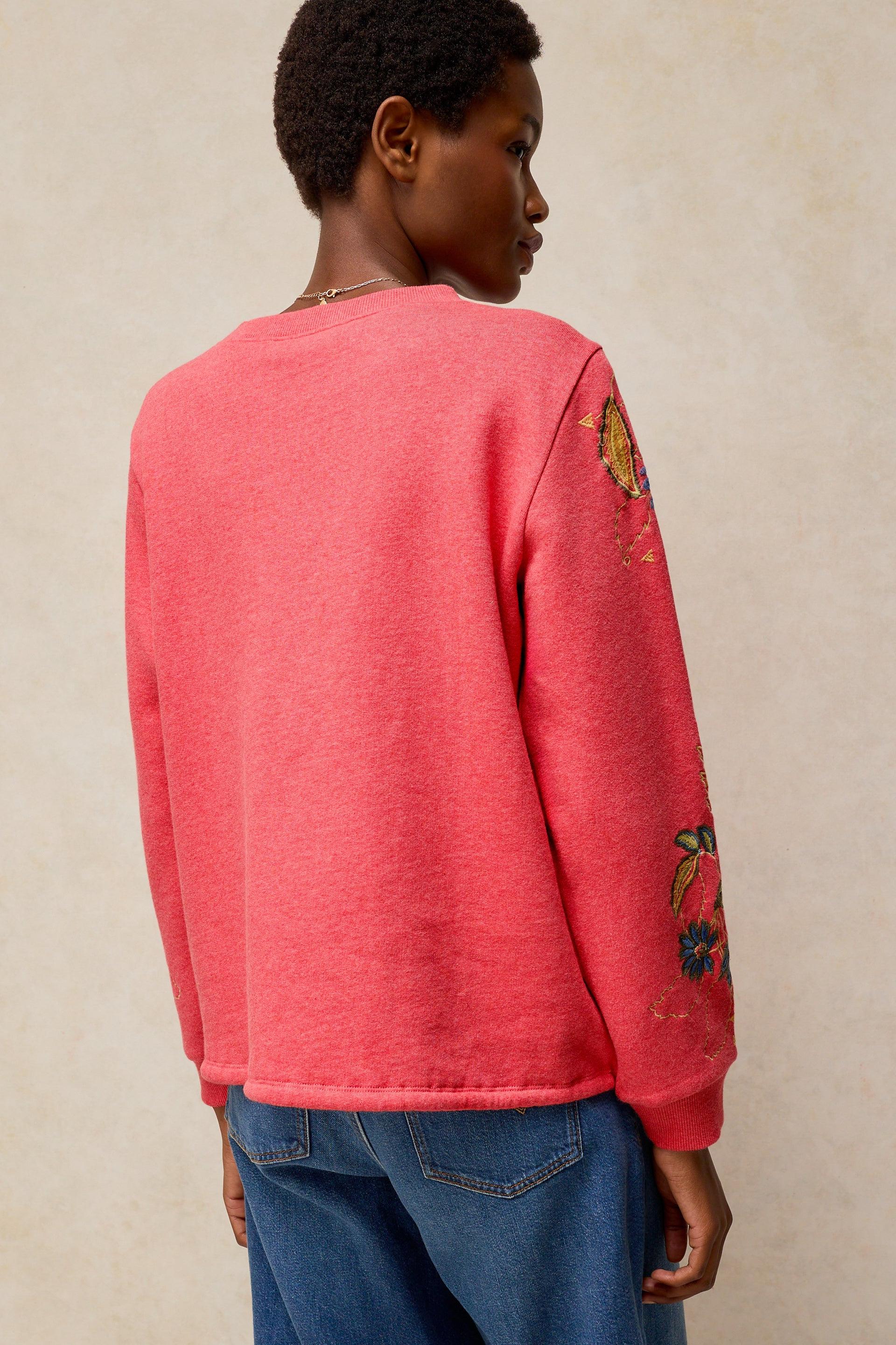 A model shown from the back wearing a coral pink crewneck sweatshirt with colorful floral embroidery on the sleeves, paired with blue jeans, against a textured beige background.