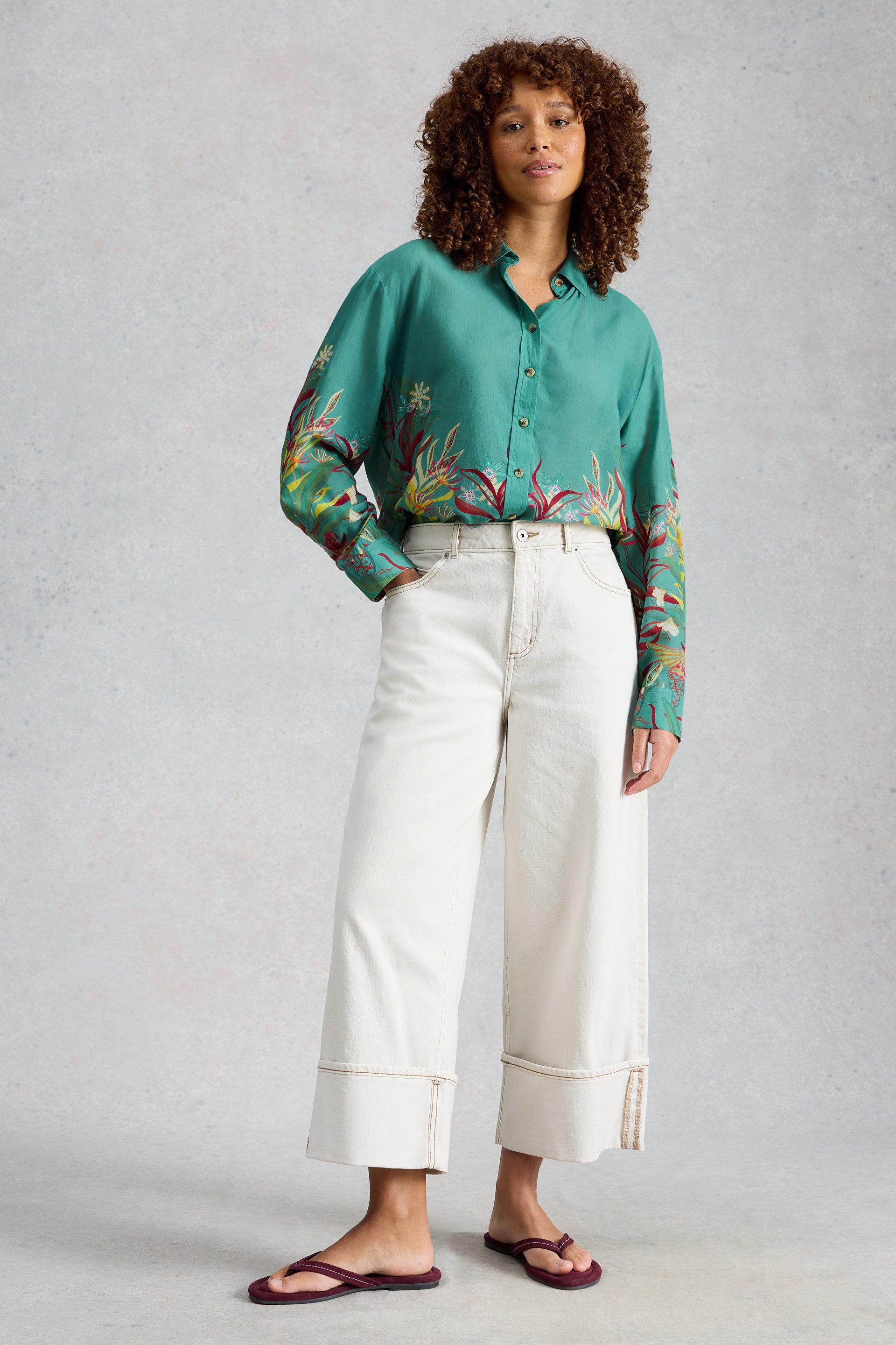 Woman wearing a green floral organic cotton printed shirt and white wide-leg trousers by White Stuff.