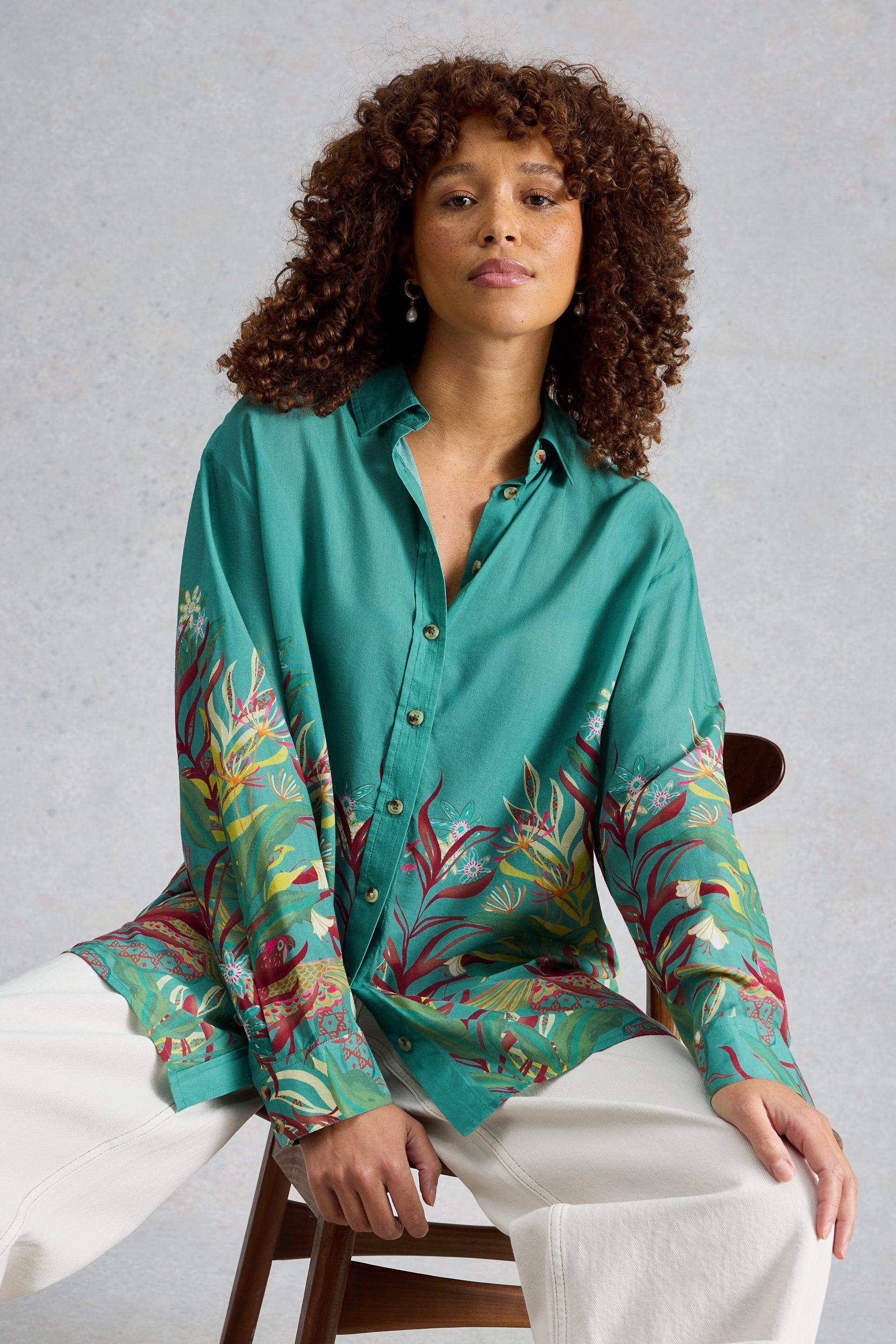 Woman with curly hair wearing a green organic cotton printed shirt sitting on a chair.