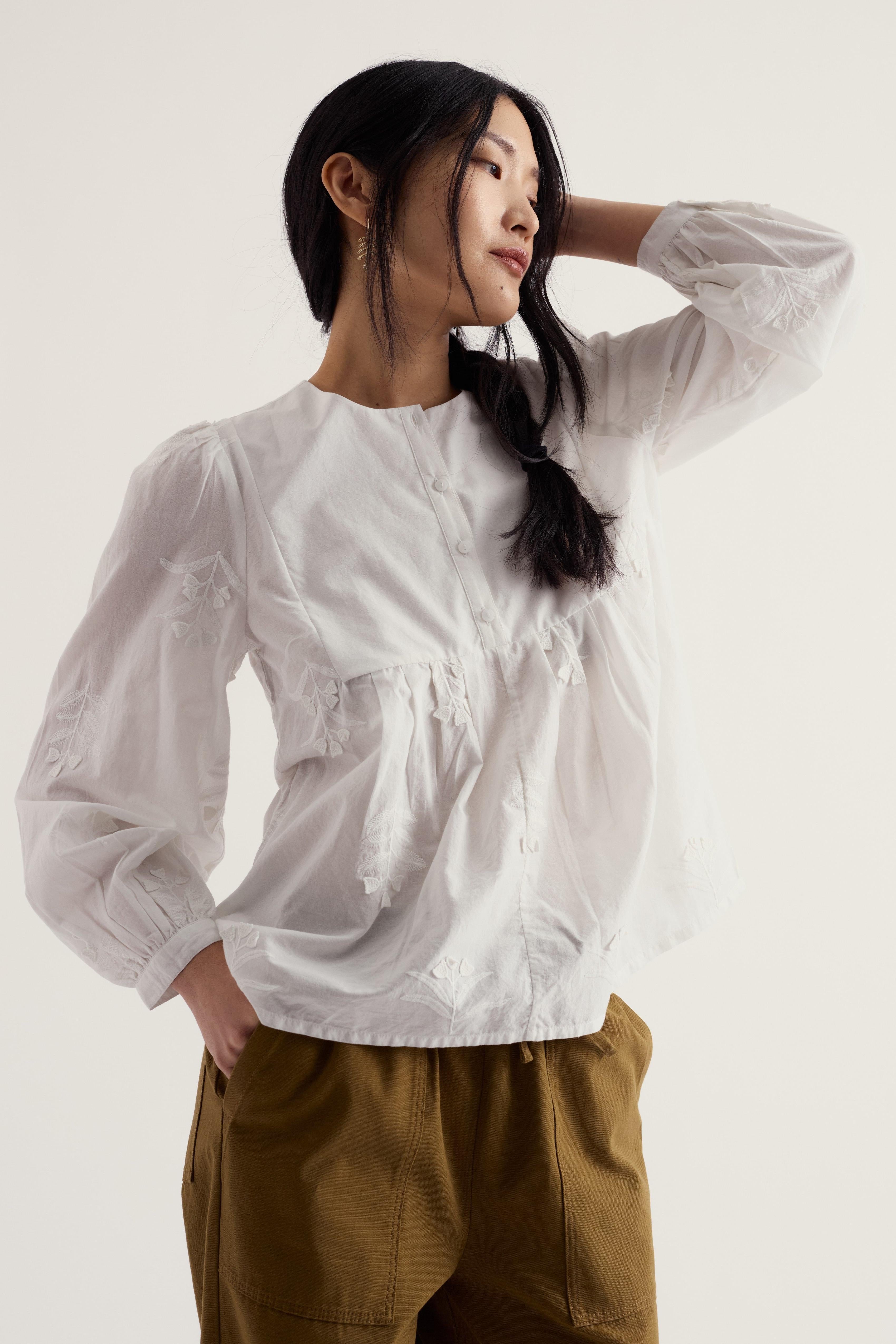 A woman wearing a white long-sleeve blouse with delicate embroidery and gathered details, paired with brown pants, photographed against a plain light background.