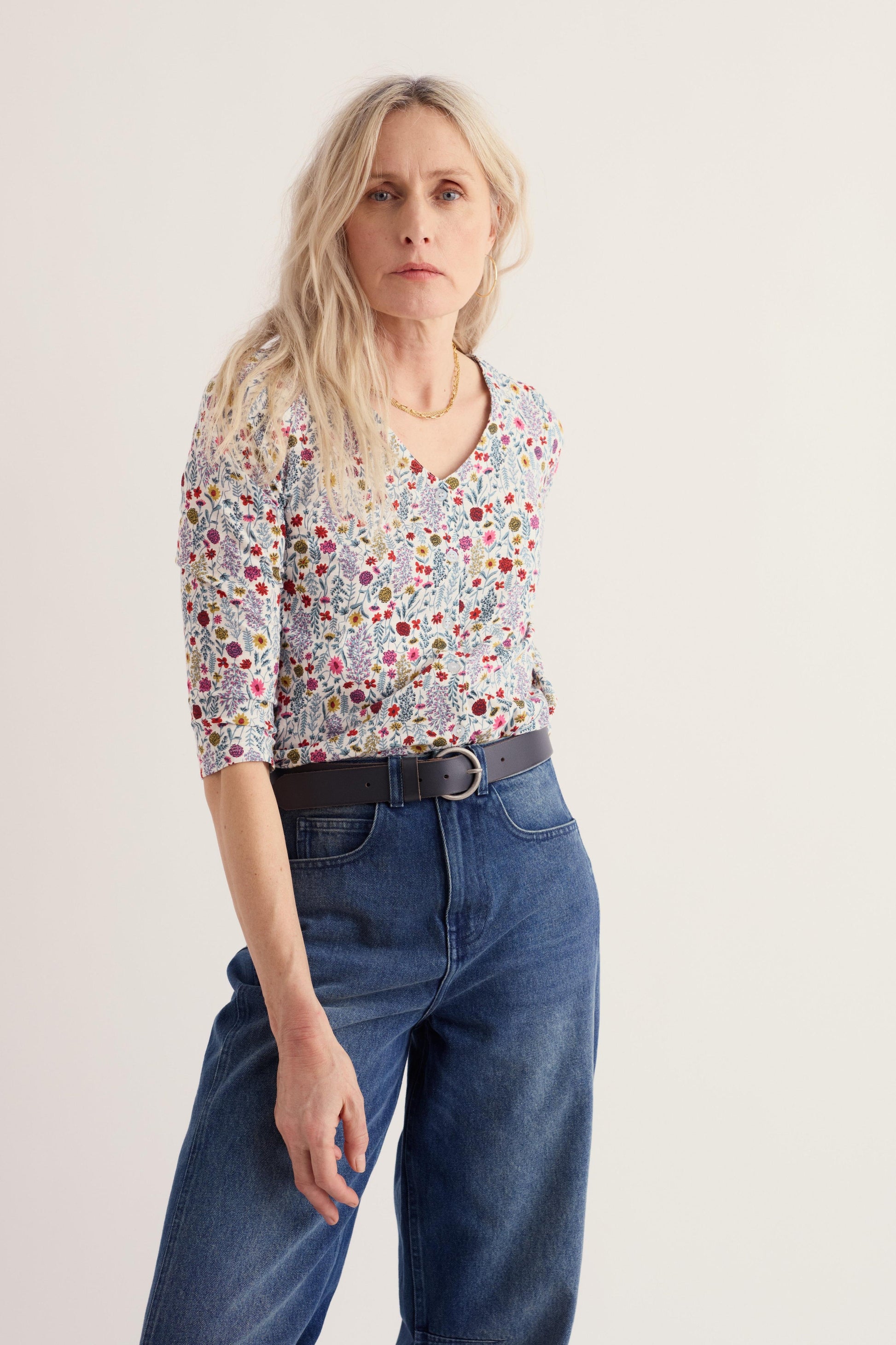 A woman modeling a short-sleeve V-neck blouse with a dense multicolor floral print, paired with a black belt and blue jeans, against a plain light background.