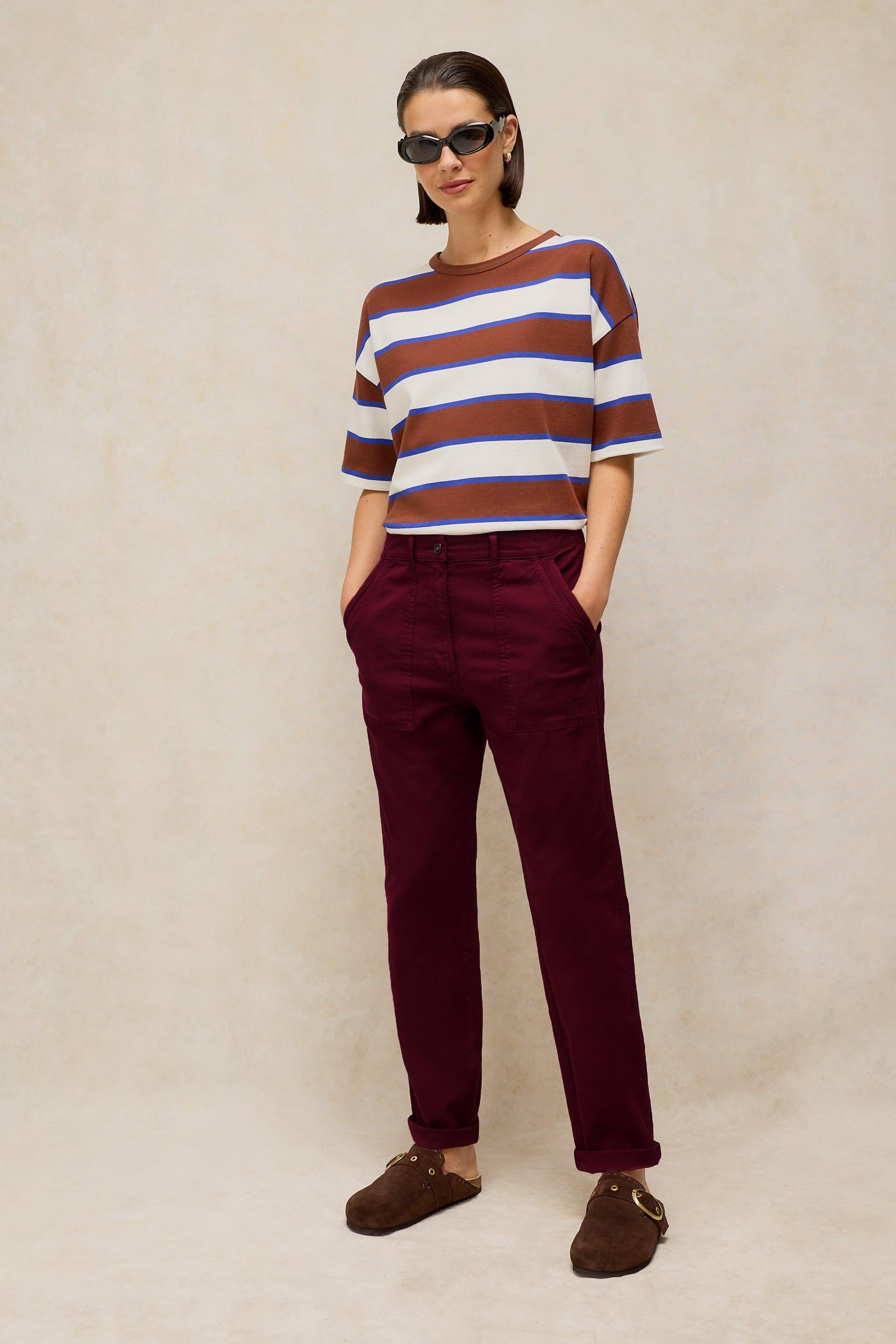 A woman wearing a striped organic cotton shirt and maroon trousers standing against a neutral studio background.