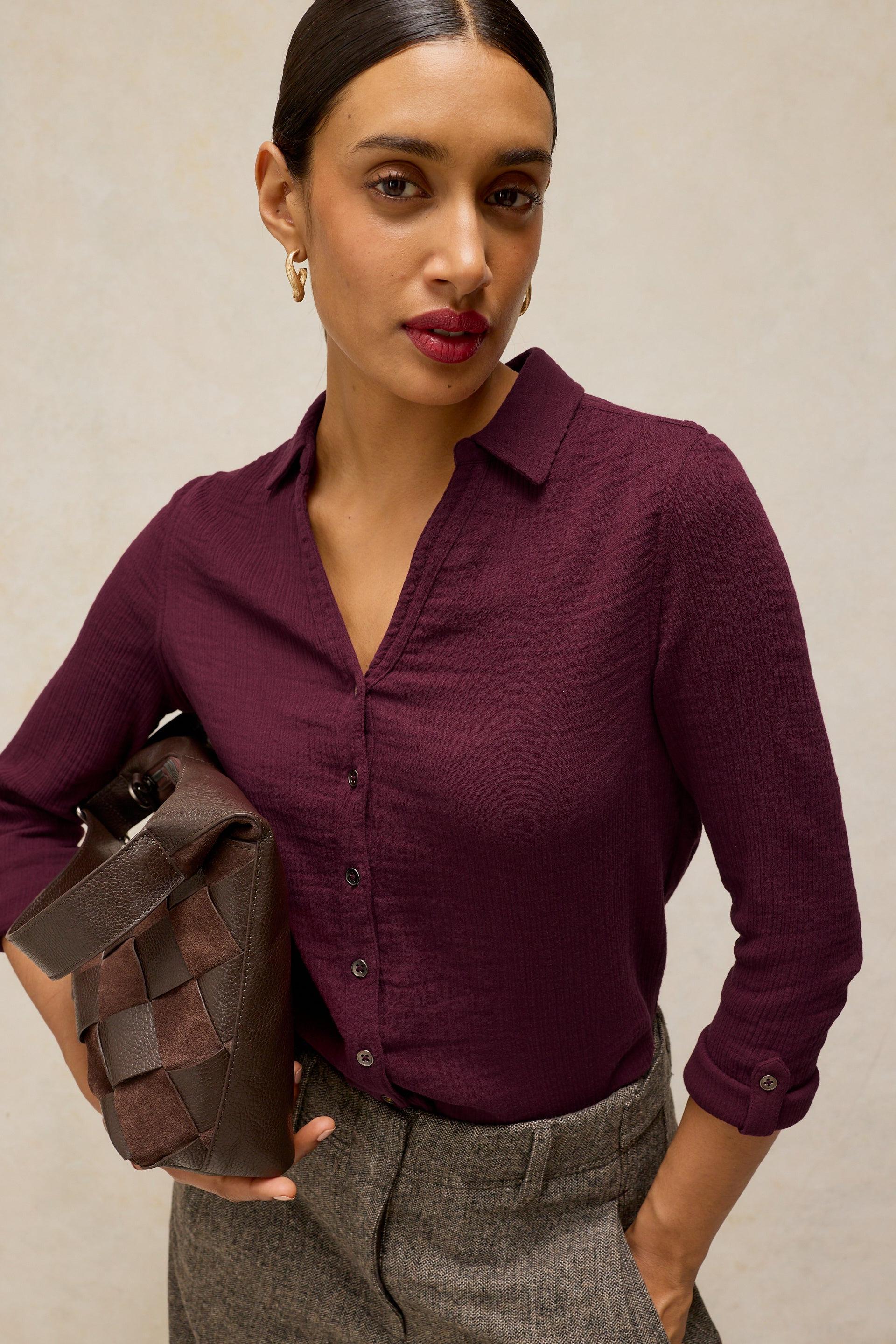 A woman wearing a long-sleeve button-up shirt in deep burgundy, paired with gray trousers and holding a dark brown woven clutch bag against a neutral beige background.