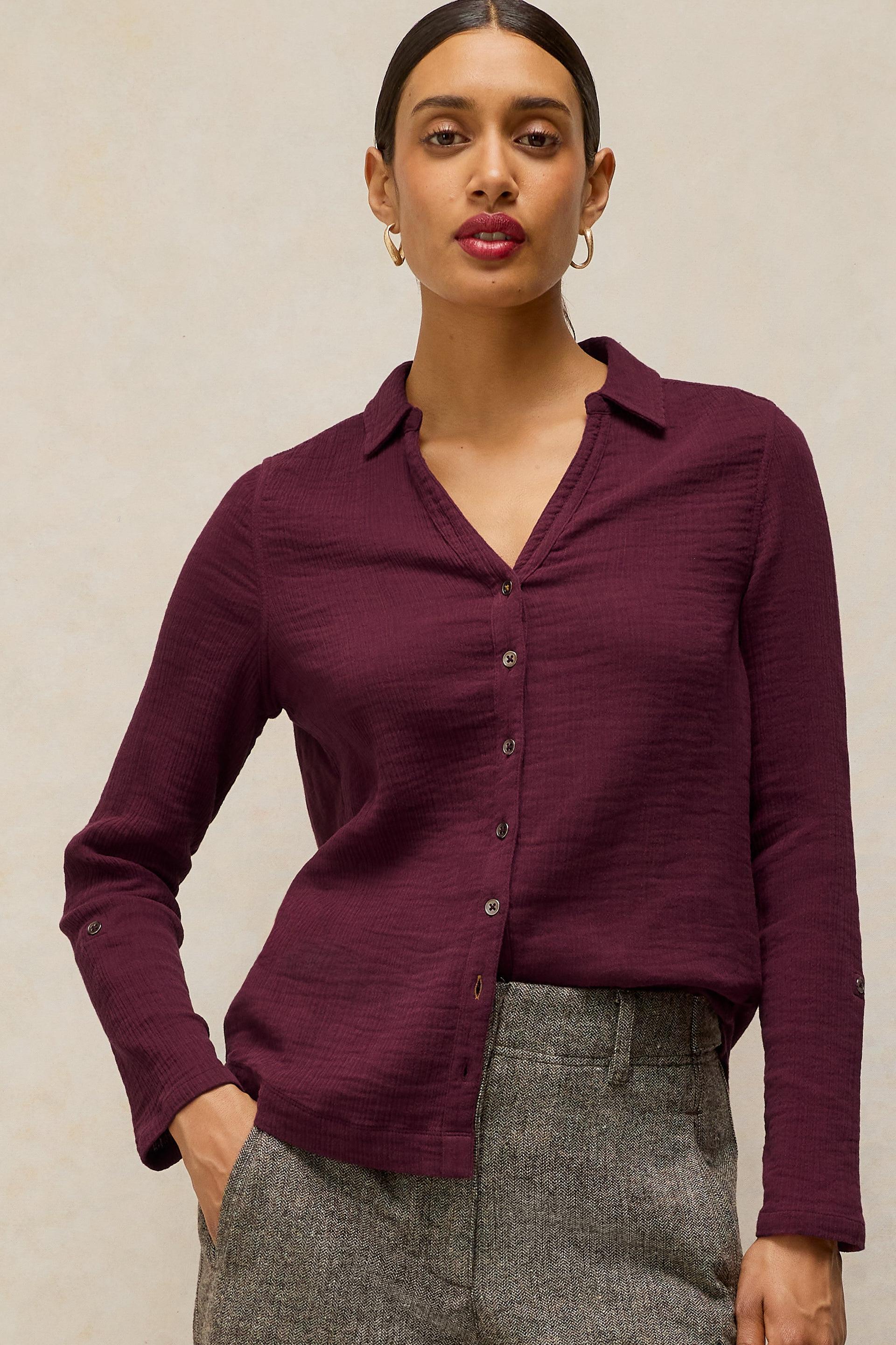 A woman wearing a long-sleeve button-front collared shirt in deep burgundy, paired with gray textured trousers, standing against a plain light beige background.