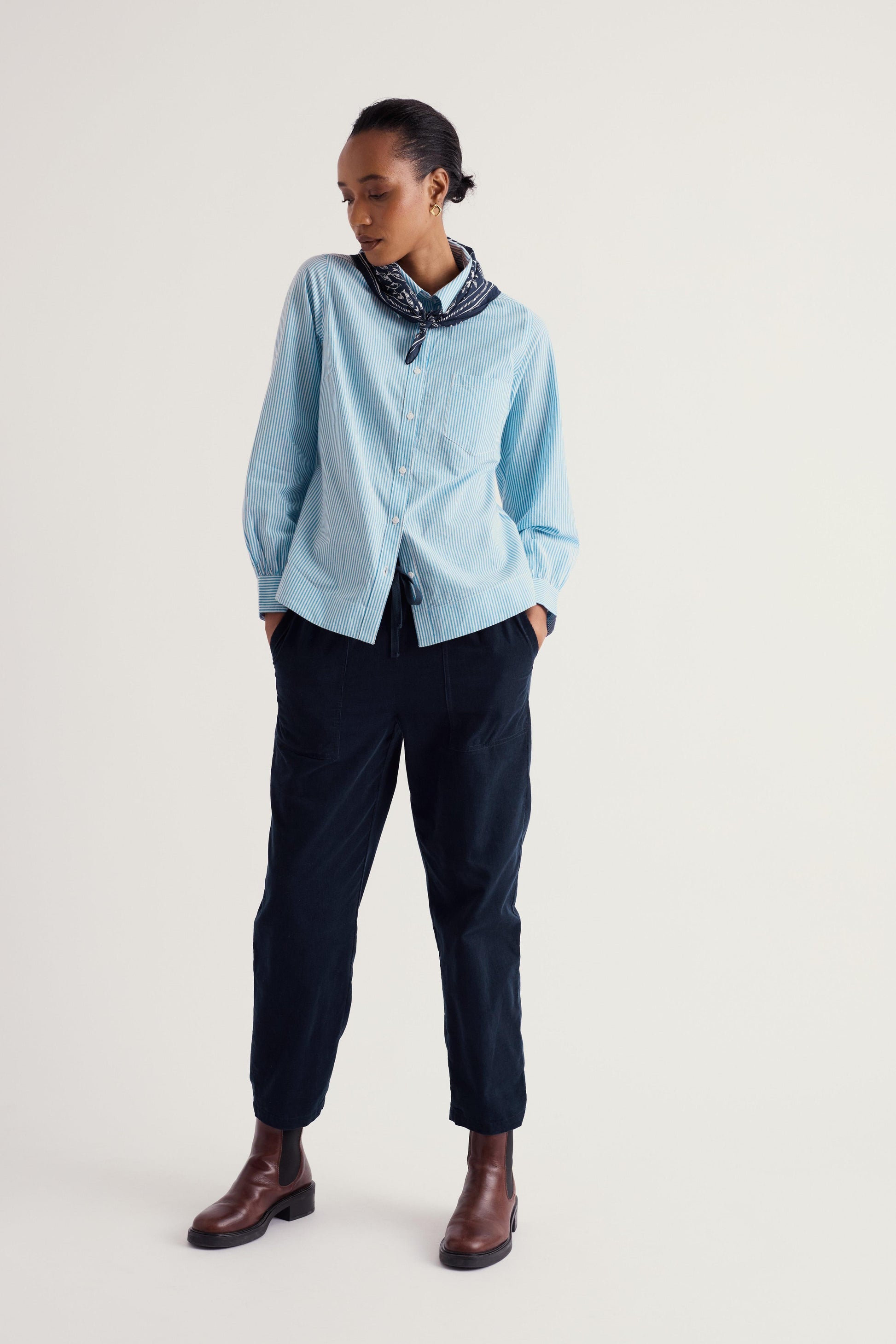 A model stands against a light gray studio background wearing a light blue long-sleeve button-up shirt, dark navy trousers, a dark patterned neck scarf, and brown leather ankle boots.