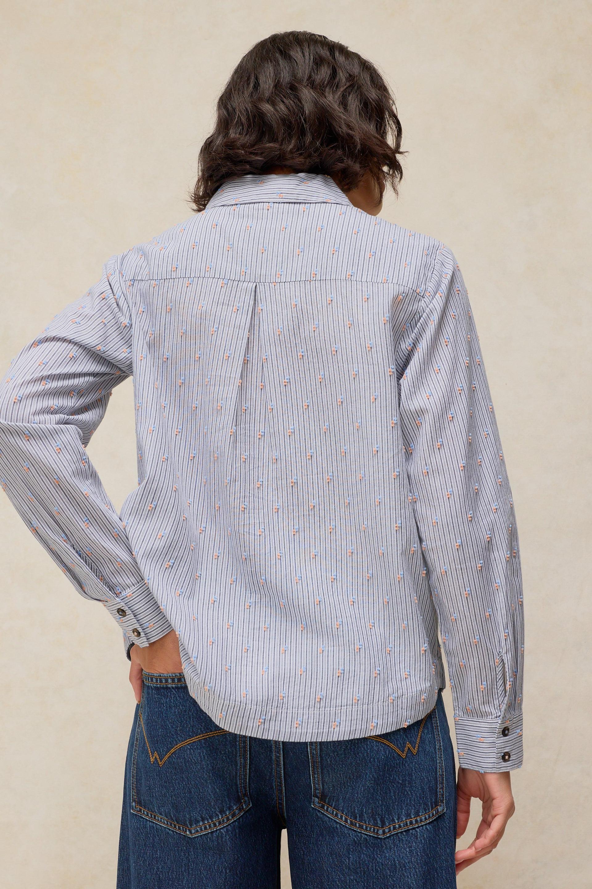 A person shown from the back wearing a light blue and white vertically striped button-up shirt with a small orange pattern, paired with blue jeans, against a textured beige background.