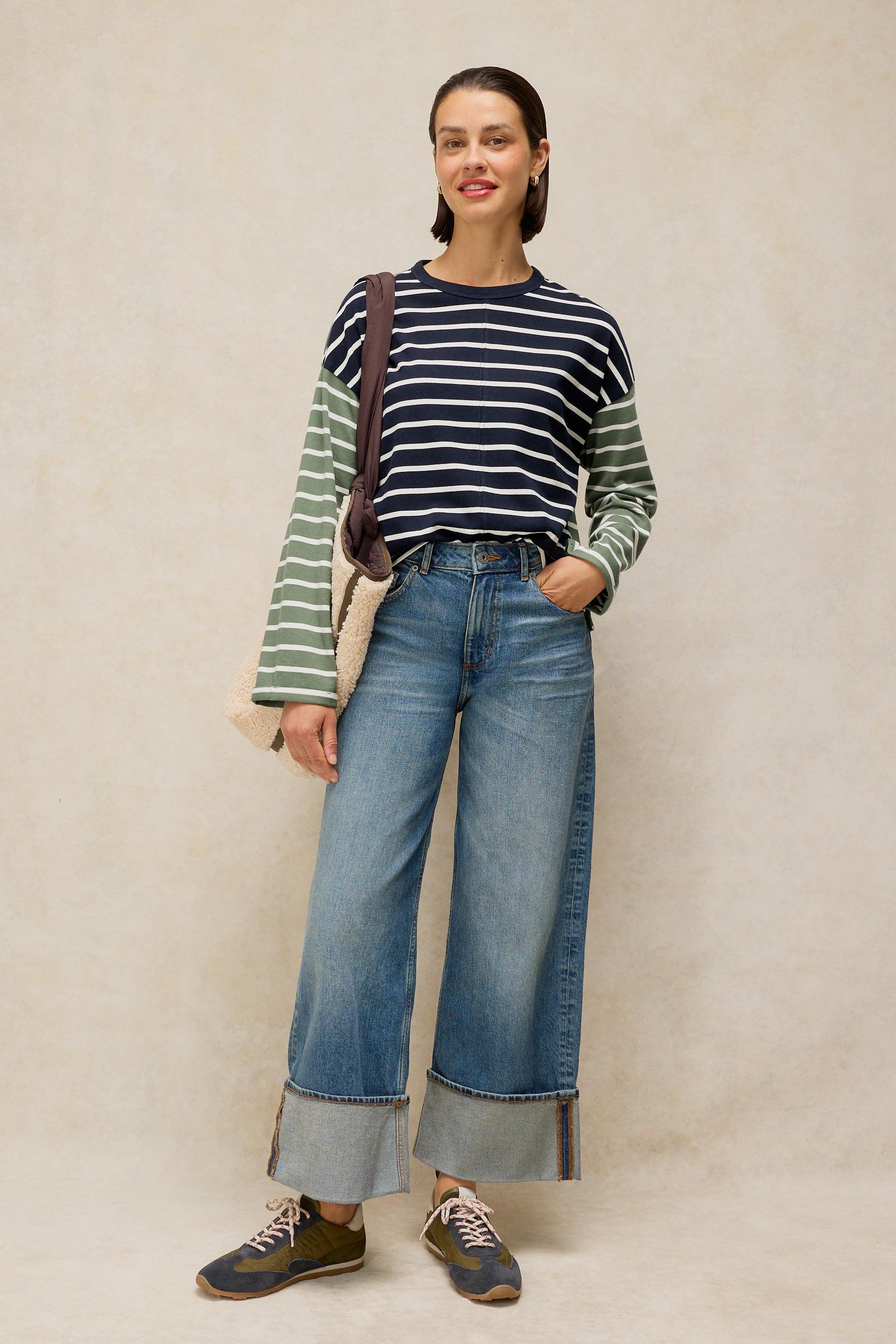 A woman stands against a textured beige studio backdrop wearing a navy and green striped long-sleeve top, wide-leg blue jeans with rolled cuffs, brown shoes, and a tan shoulder bag.