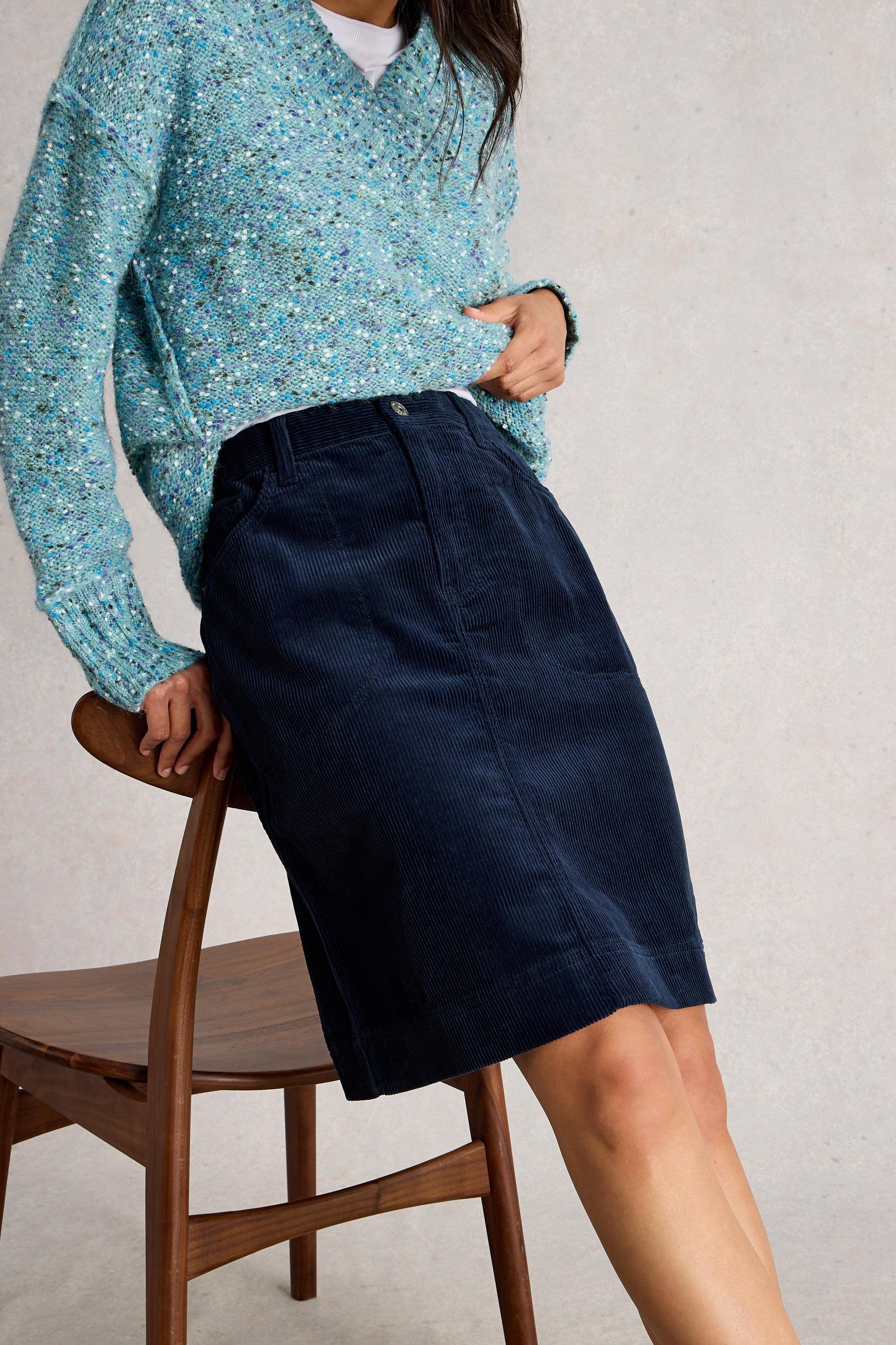 A model wearing a textured light blue V-neck sweater and a dark navy blue corduroy skirt, sitting on a wooden chair against a neutral light gray background.