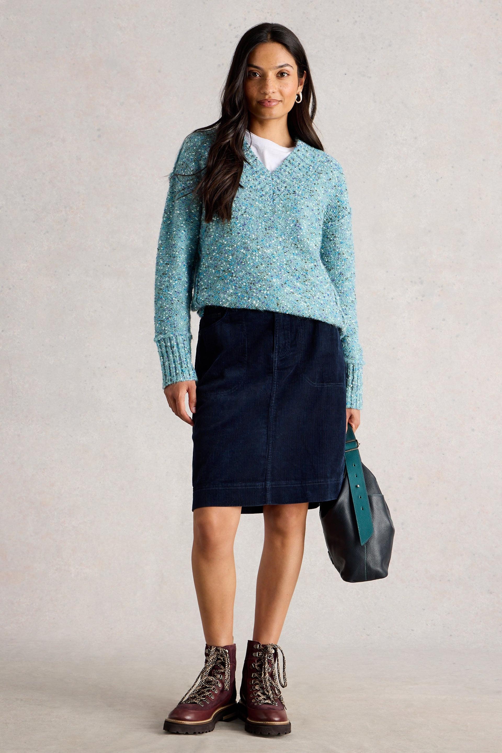 Woman wearing a light blue sweater, dark blue skirt, and brown boots against a plain background