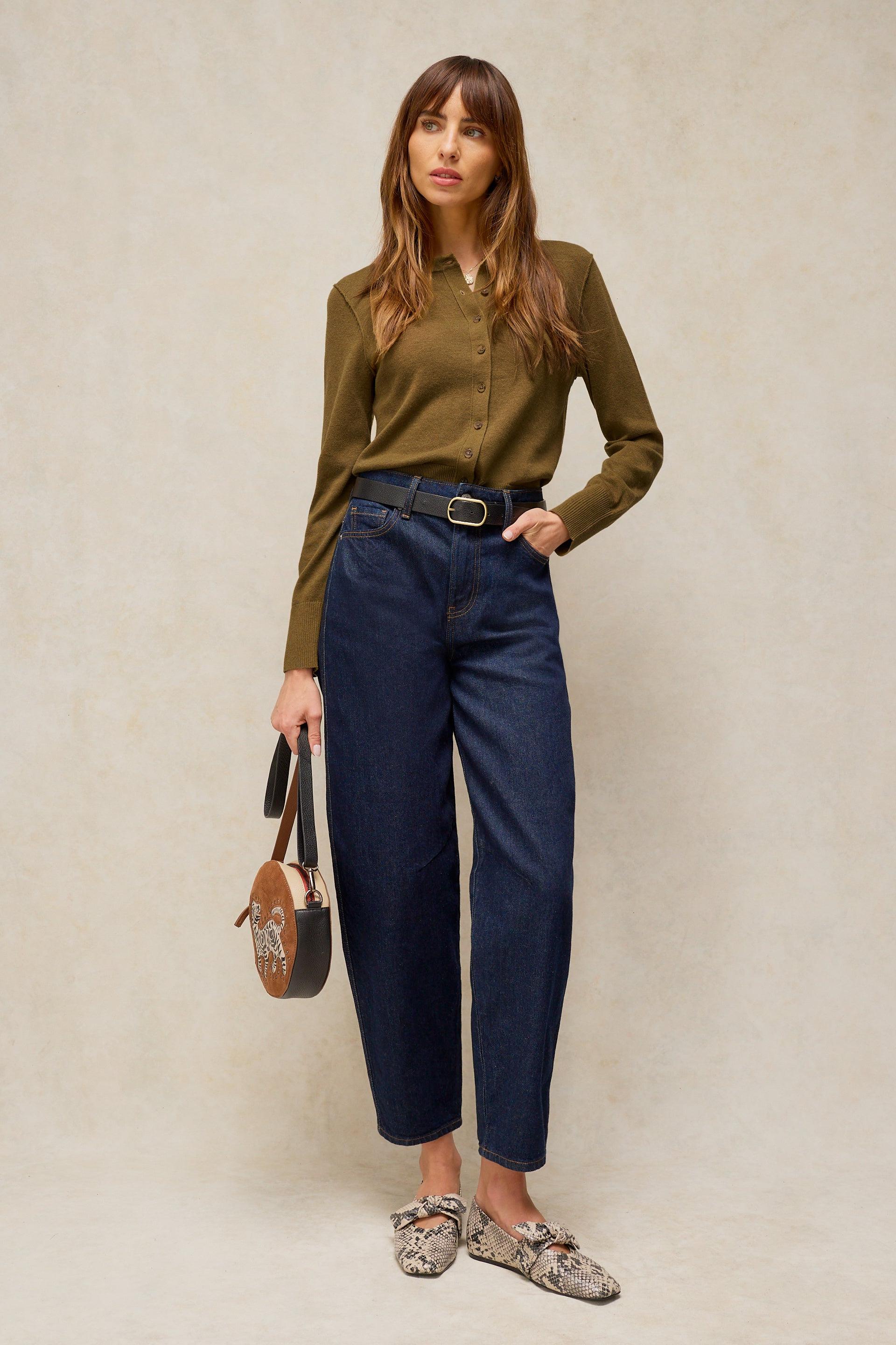 A woman stands against a textured beige studio backdrop wearing an olive green long-sleeve button-up cardigan, dark blue cropped jeans with a black belt, and patterned loafers. She is holding a small brown handbag.