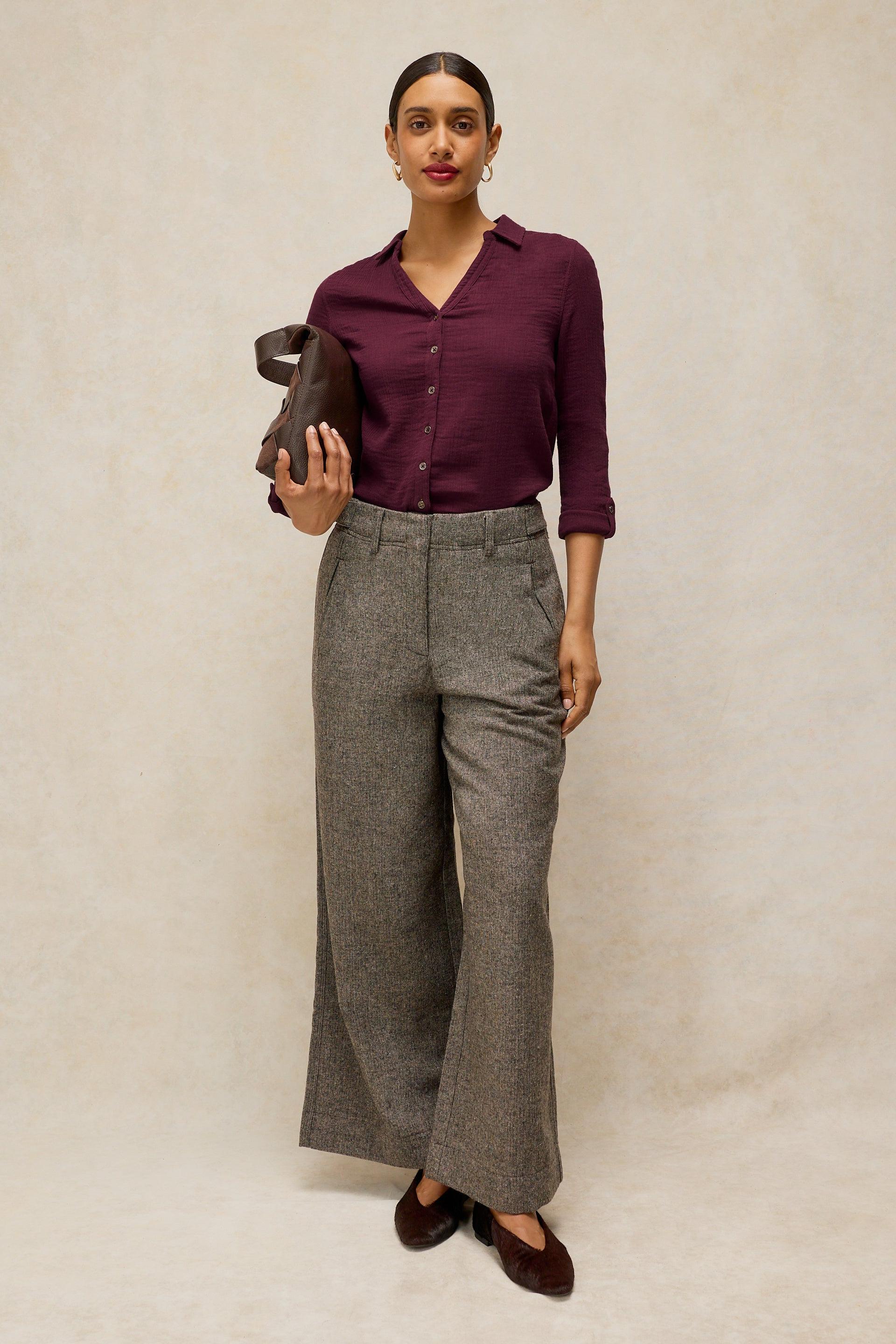 A woman stands against a textured beige studio backdrop wearing a dark purple button-up shirt, high-waisted wide-leg gray trousers, and dark brown flats, while holding a dark brown leather clutch.