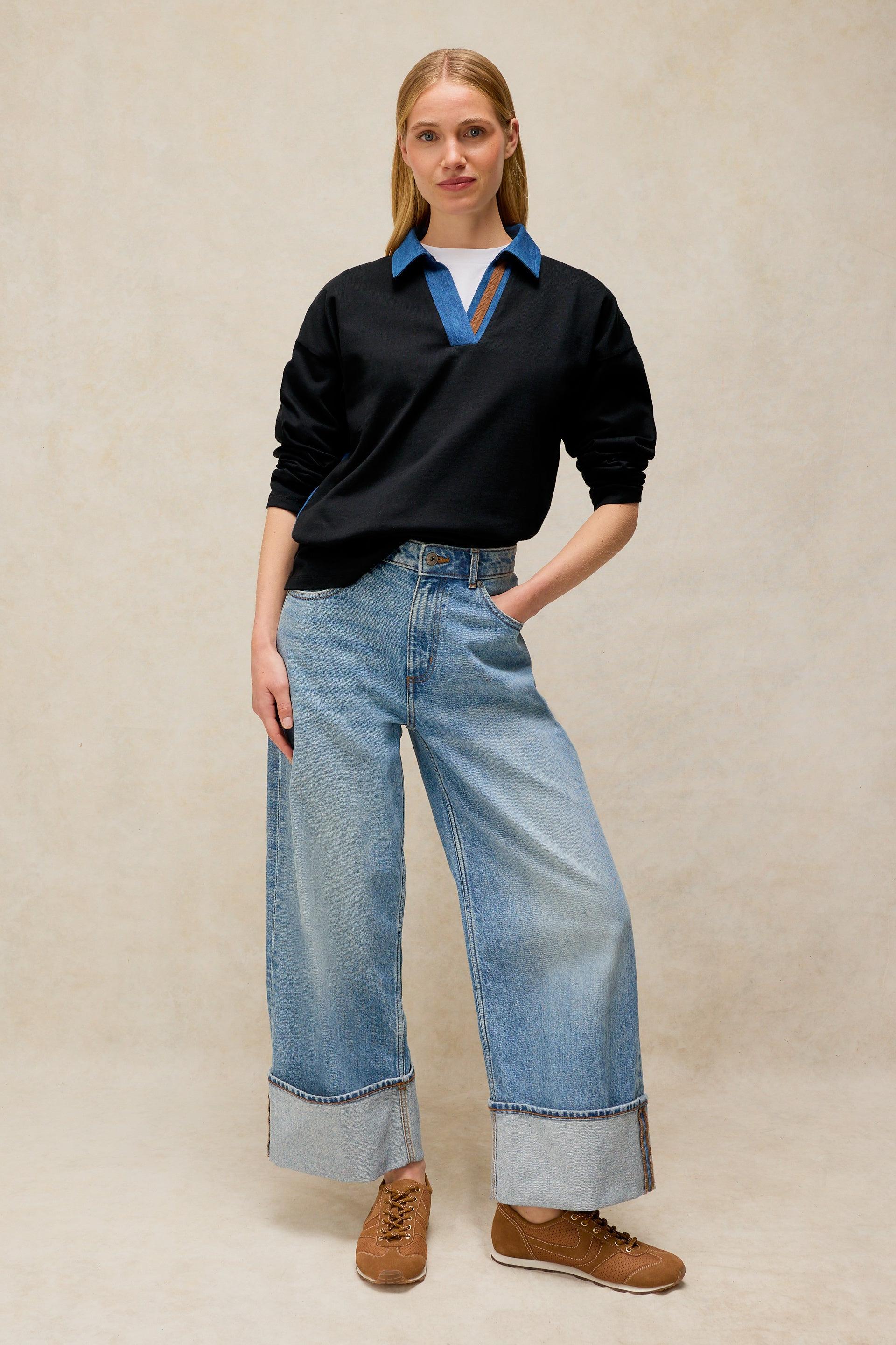 A woman posing in a black polo top layered over a white organic cotton shirt with wide-leg blue jeans