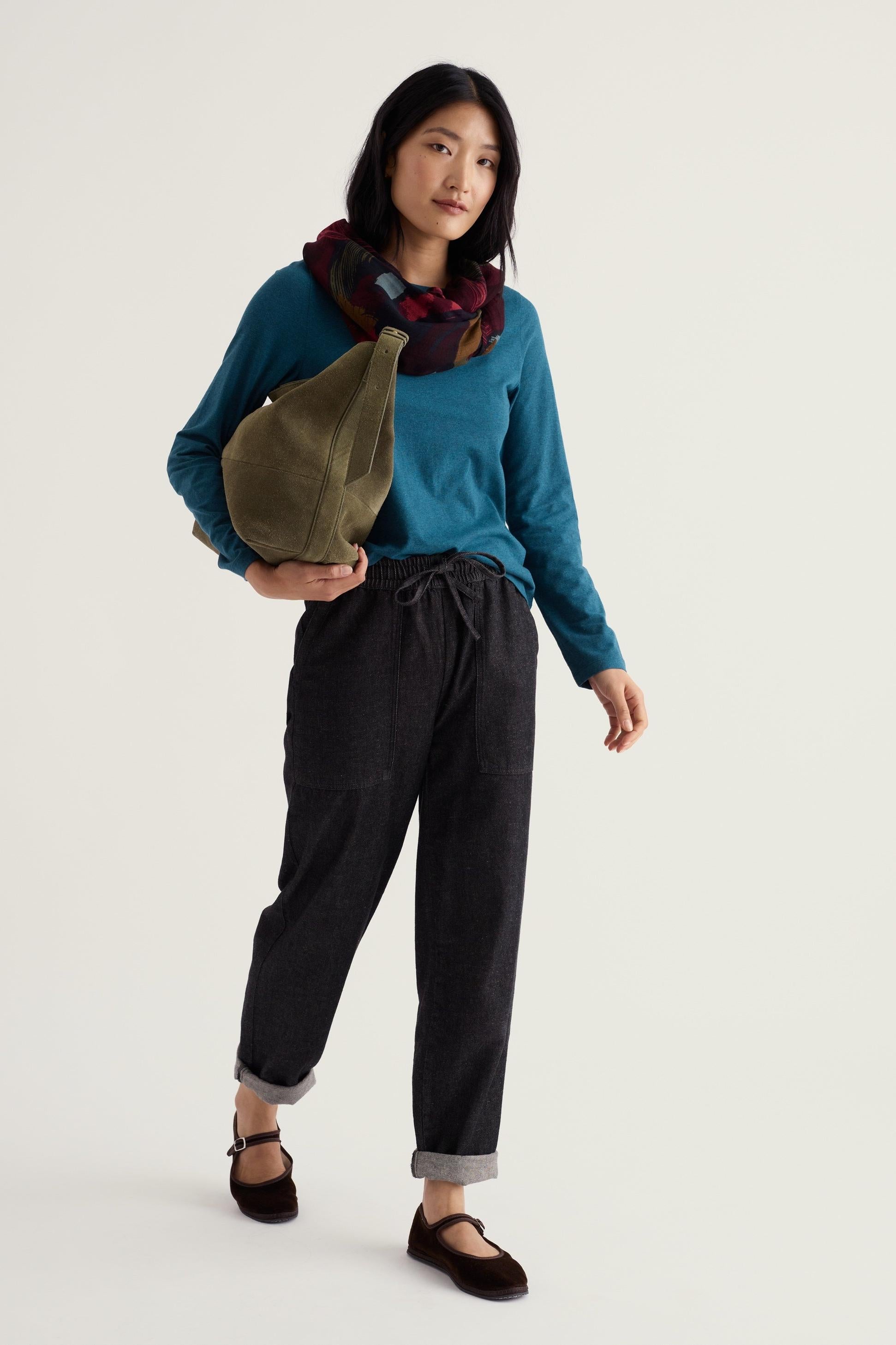 A woman posing in a blue long-sleeve organic cotton top and dark trousers.