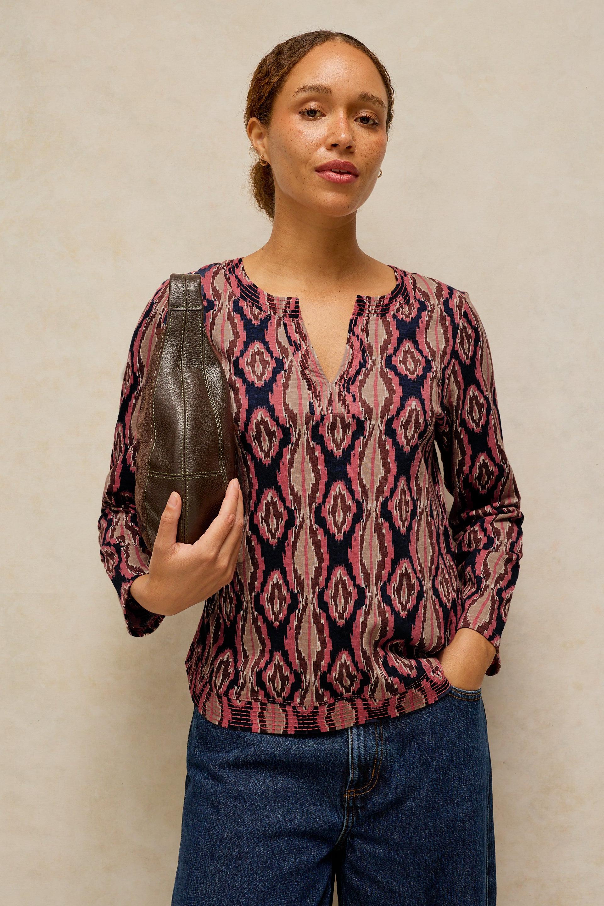 A woman wearing a long-sleeve knit top with a pink, red, and navy ikat pattern, paired with blue jeans, holding a dark brown leather shoulder bag against a textured beige wall.