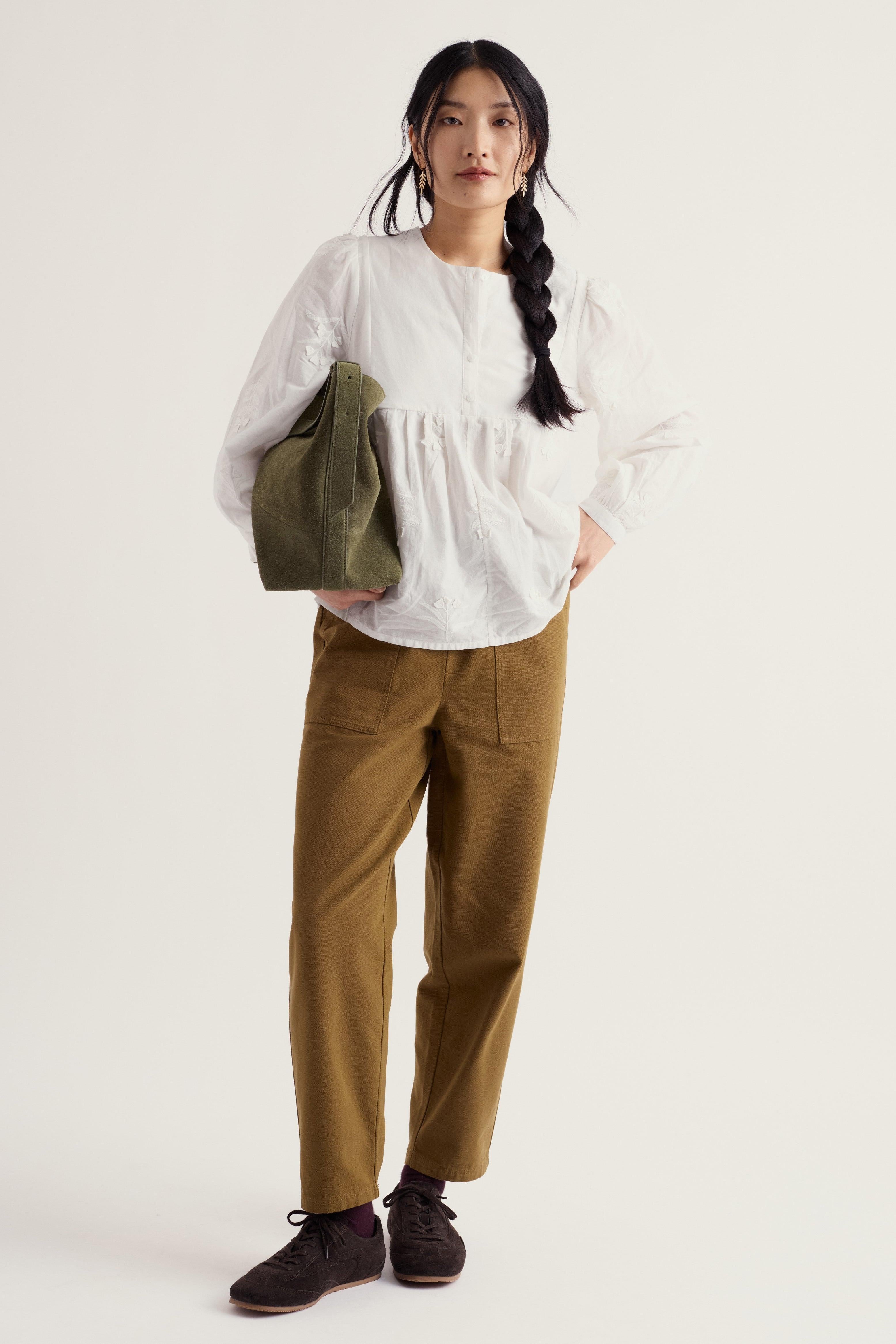 A woman stands against a plain light background wearing a loose white long-sleeve blouse, brown pants, and dark shoes, while carrying an olive green shoulder bag.