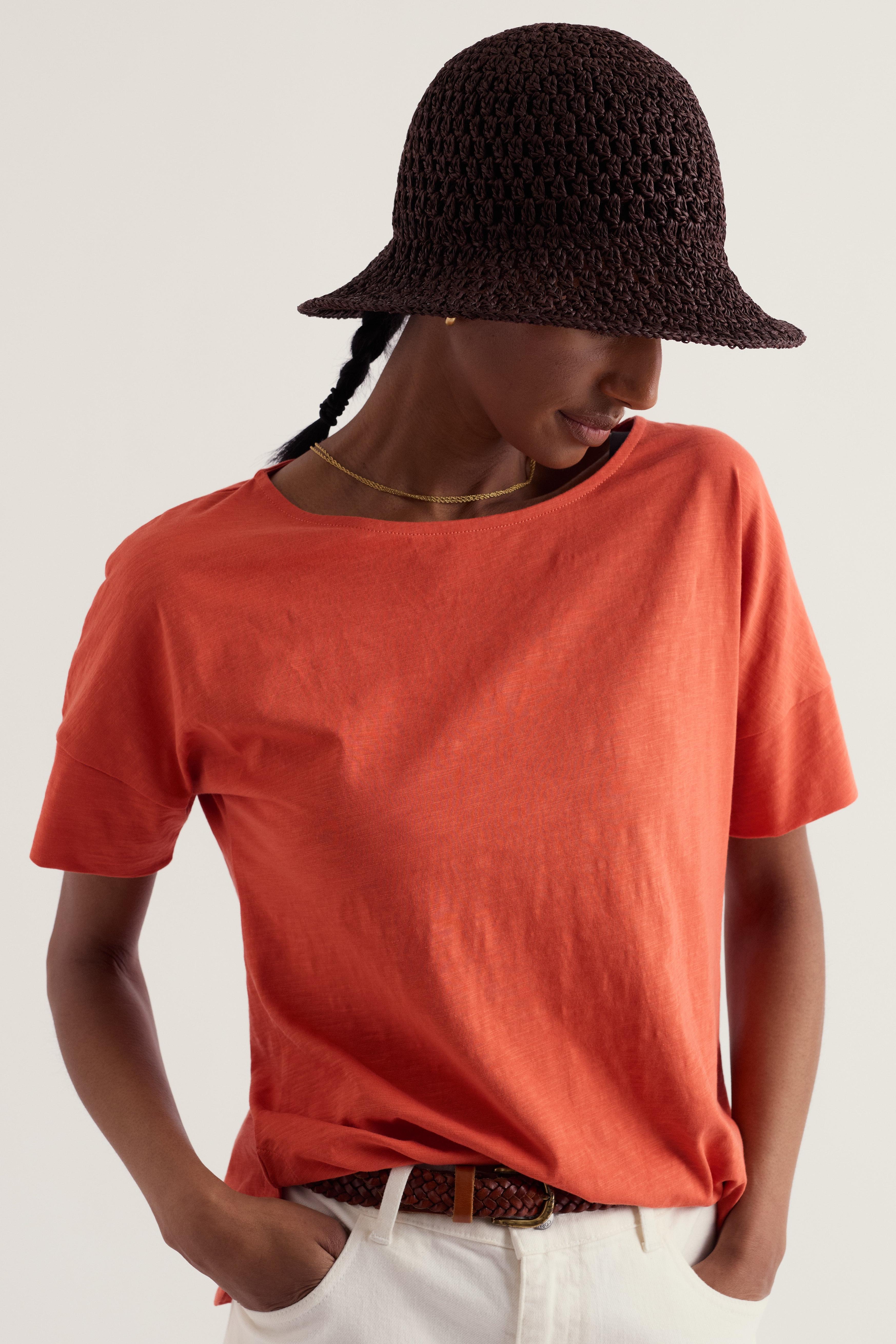 Woman wearing an orange organic cotton Seasalt t-shirt and a dark brown crochet bucket hat.
