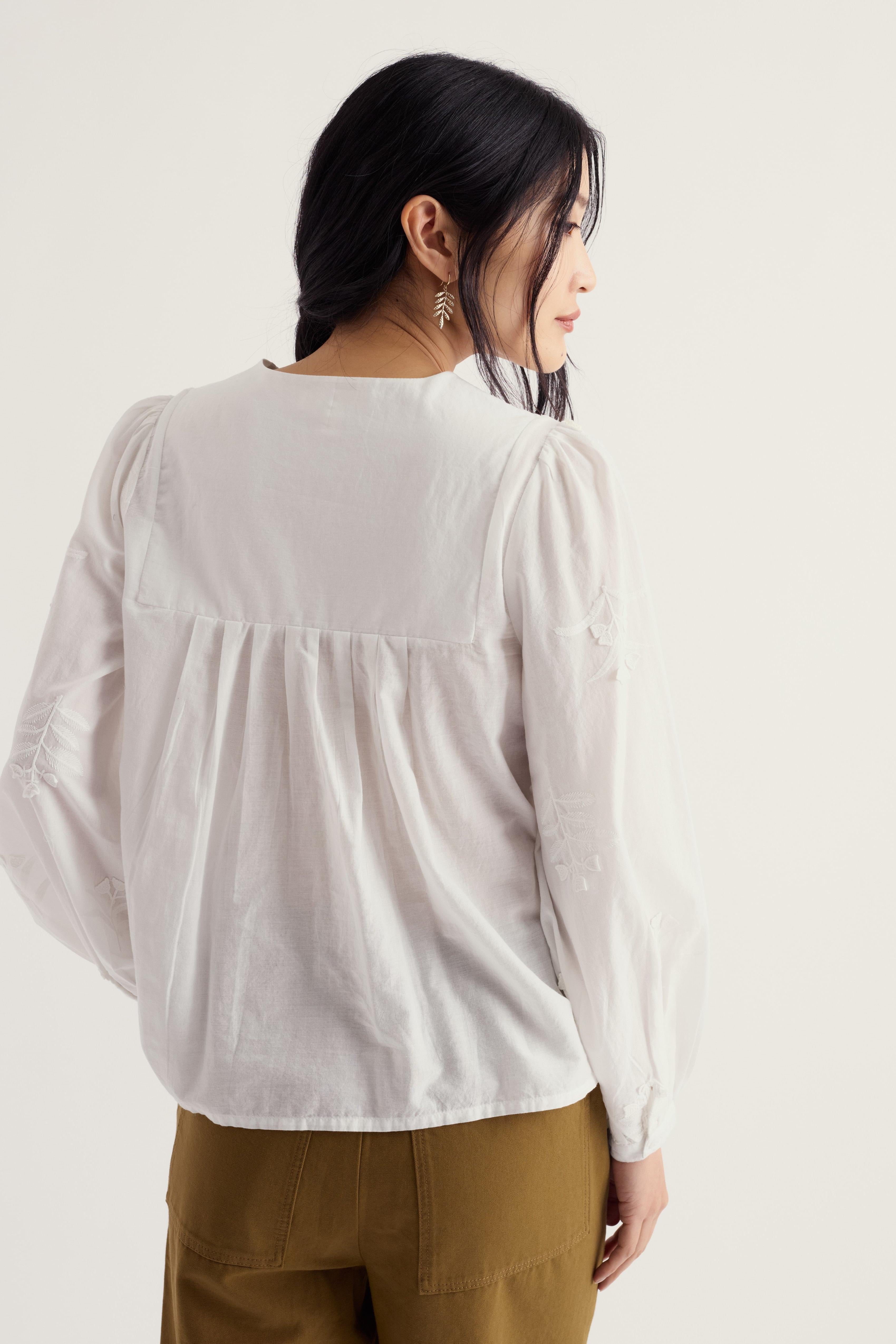 A woman shown from the back wearing a white long-sleeve blouse with a gathered, ruffled yoke and brown pants, photographed against a plain light background.
