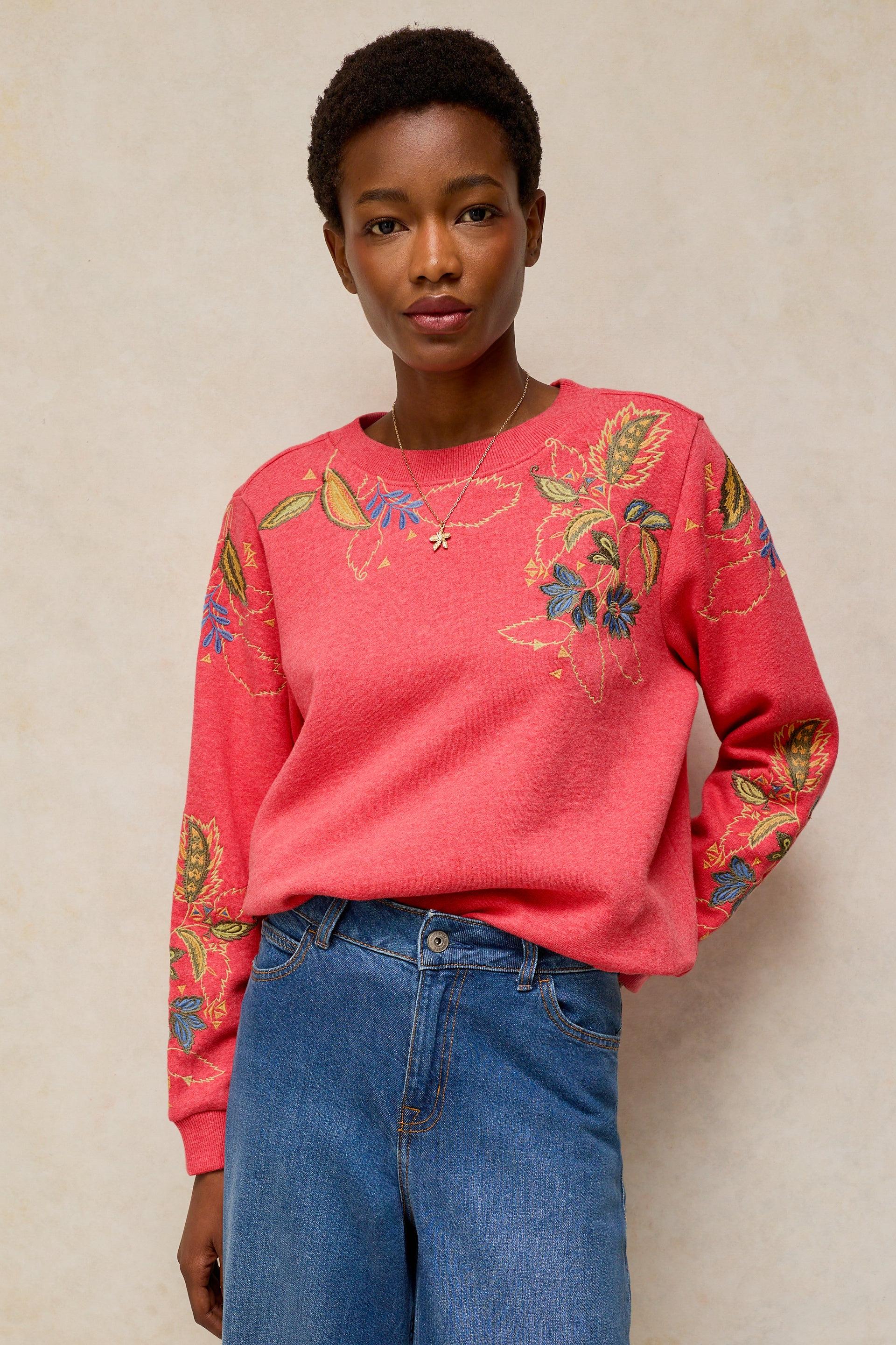 A model wearing a coral pink crewneck sweatshirt with colorful floral embroidery on the front and sleeves, paired with blue jeans, set against a neutral beige background.