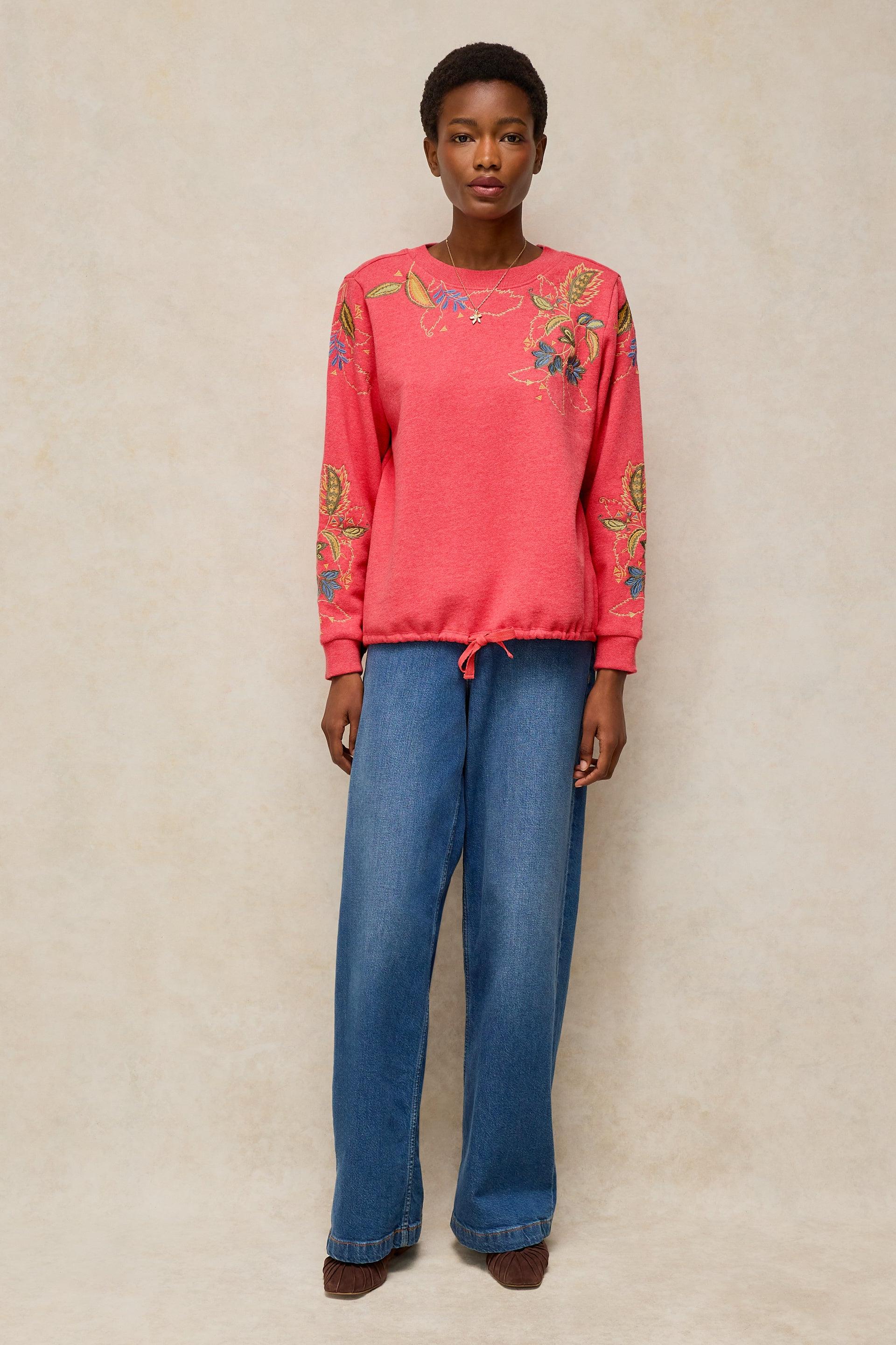 A model stands against a textured beige studio backdrop wearing a coral pink crewneck sweatshirt with colorful floral embroidery on the chest and sleeves, paired with wide-leg blue jeans.