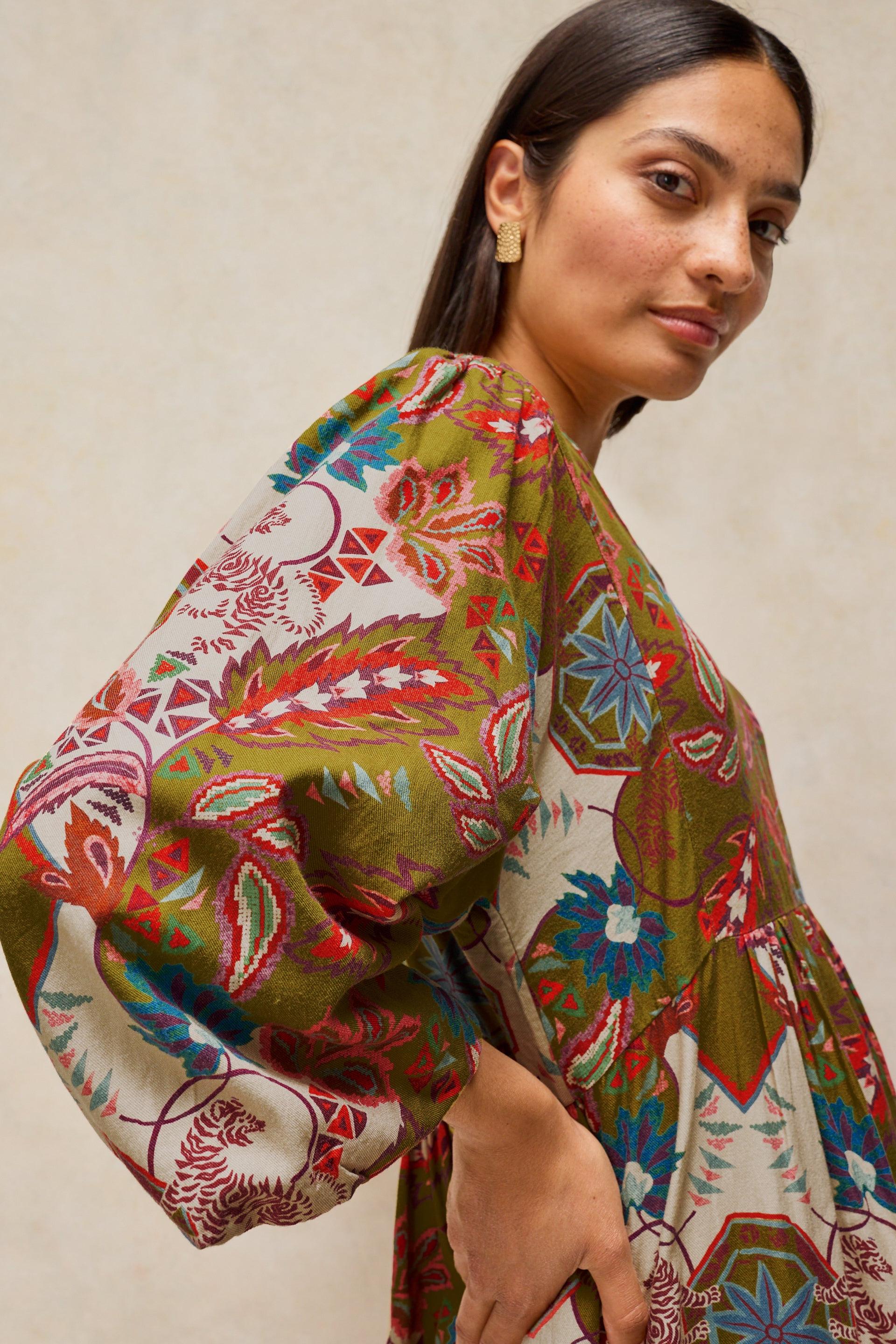 A woman wearing a loose, long-sleeve dress with a vibrant multicolor floral and geometric print in olive green, red, blue, and cream, set against a neutral beige background.