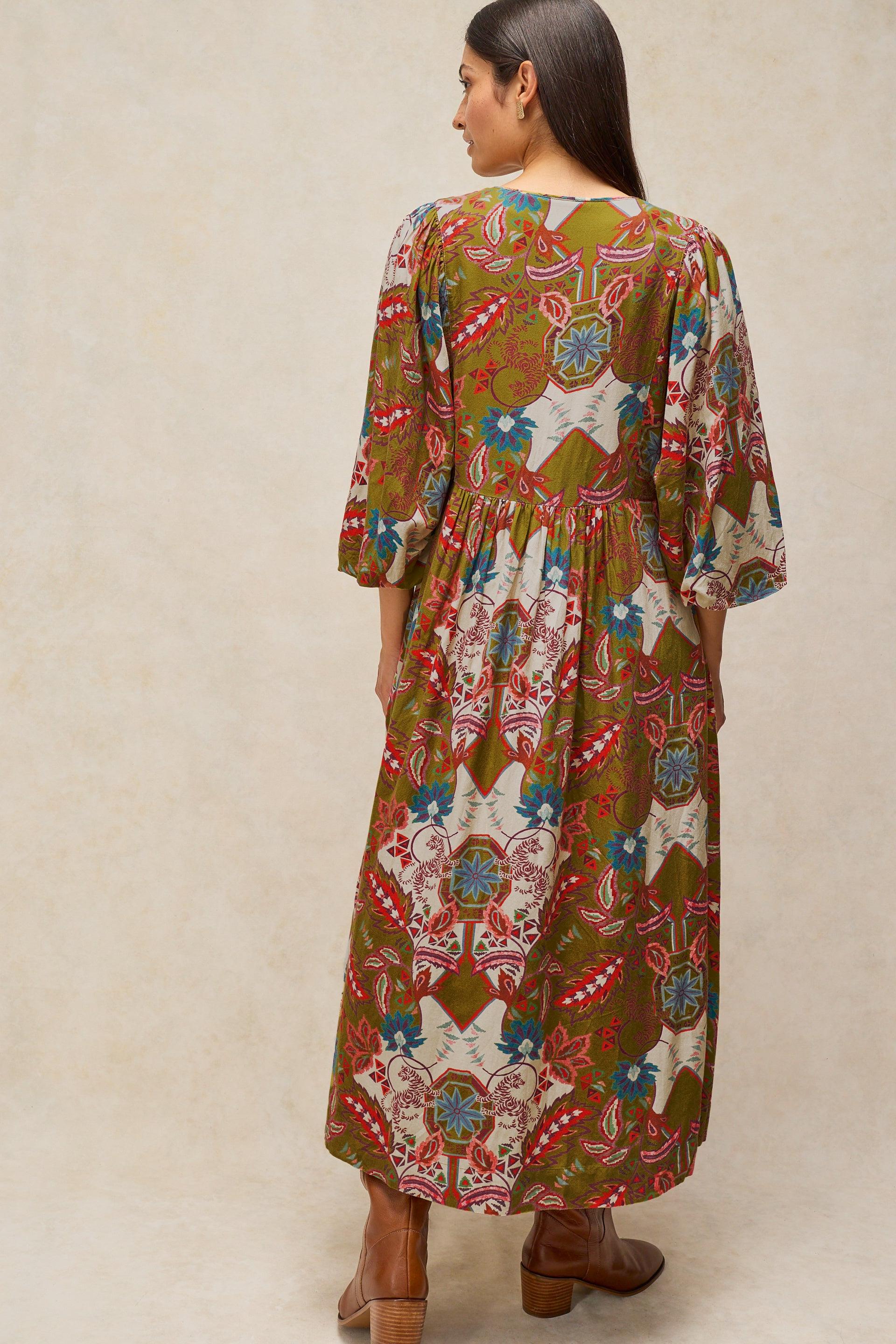 A woman shown from the back wearing a long-sleeved, knee-length kaftan dress with a colorful geometric and floral pattern on an olive green base, paired with brown leather ankle boots, set against a neutral beige background.